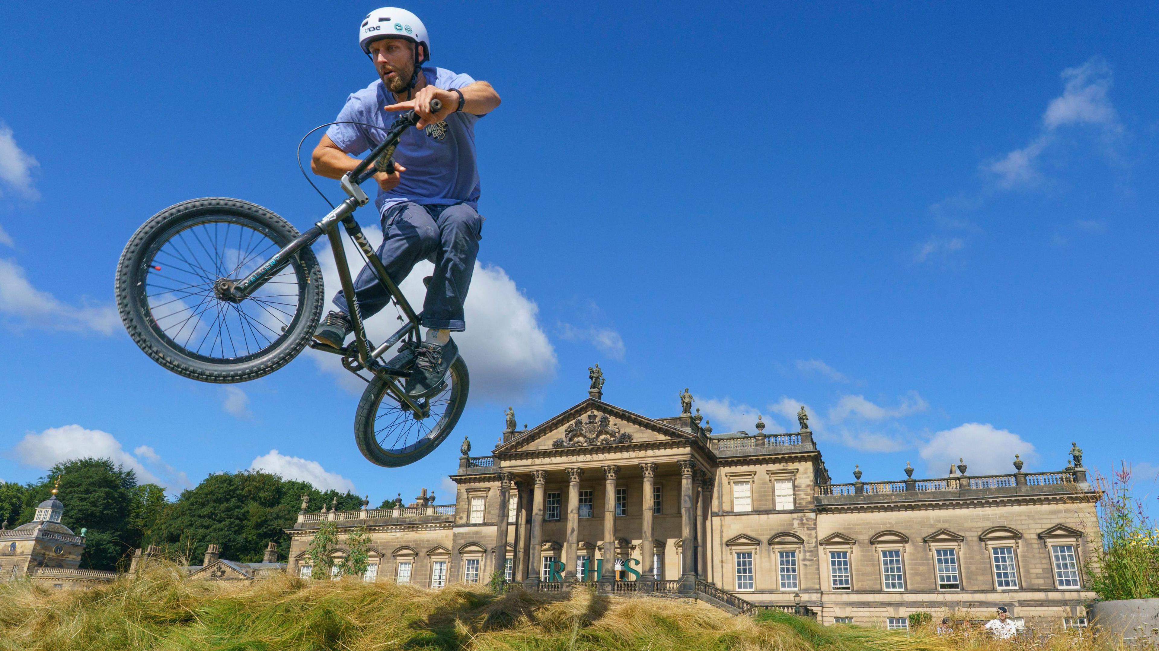 The photo is taken from below showing a cyclist fly through the air on a small bike. He is wearing jeans, a blue t shirt and white helmet. The stately home is in the background. It is a grand, sandstone building with rows of windows at the front and pillars ahead of the main entrance.