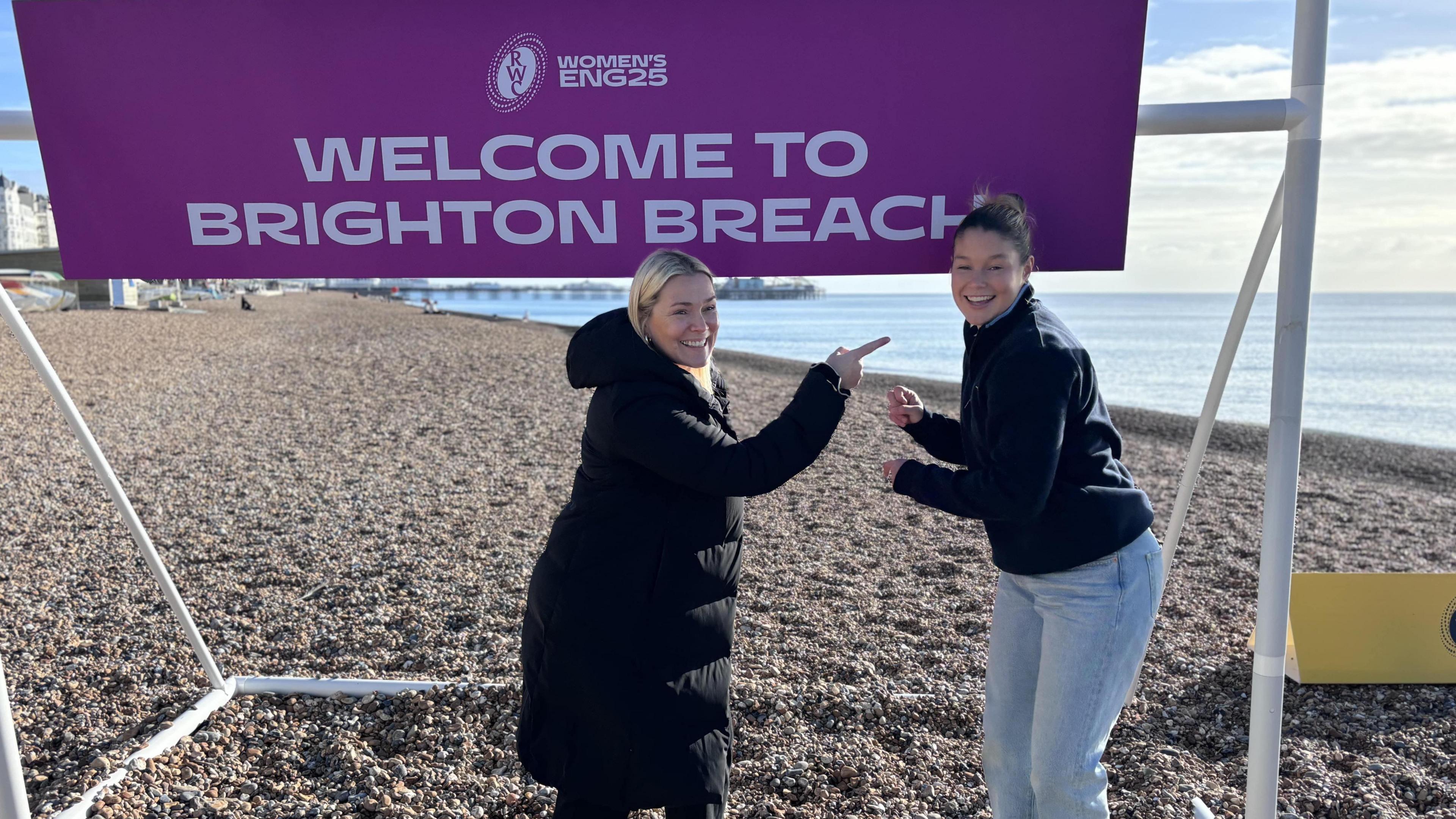 Jess Breach (right, a woman with light blue jeans, a dark blue jumper and her hair in a bun, smiling looking into the camera) and Rachael Burford (left, a woman with blonde hair and a long black coat pointing a Jess) standing in front of a purple sign with white writing on saying Welcome To Brighton Breach on Brighton beach