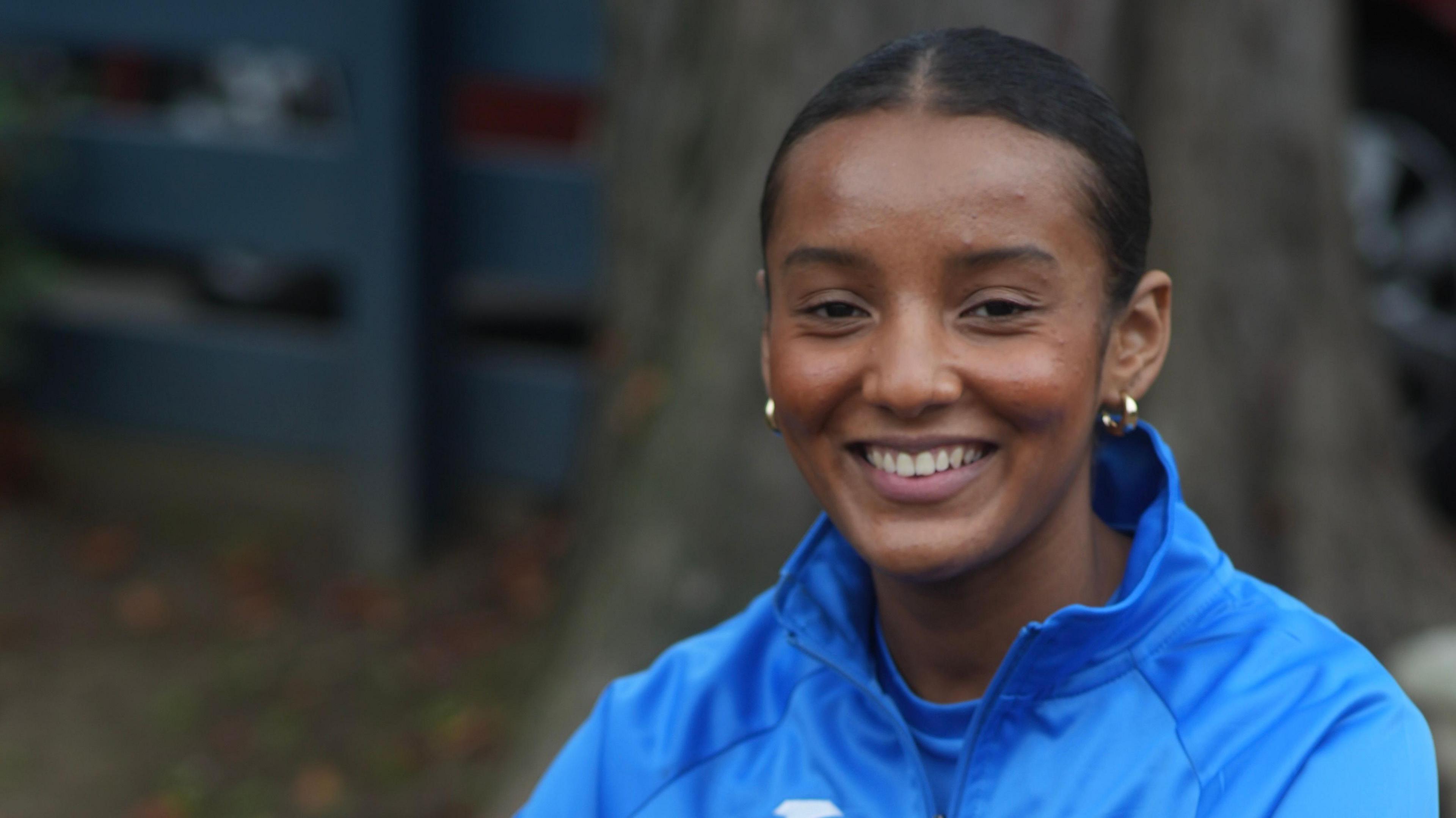 Justice Hall is smiling at the camera while sitting down. Her dark hair is tied up behind her head. She is wearing gold hoop earrings and a blue jumper with a blue top on underneath.