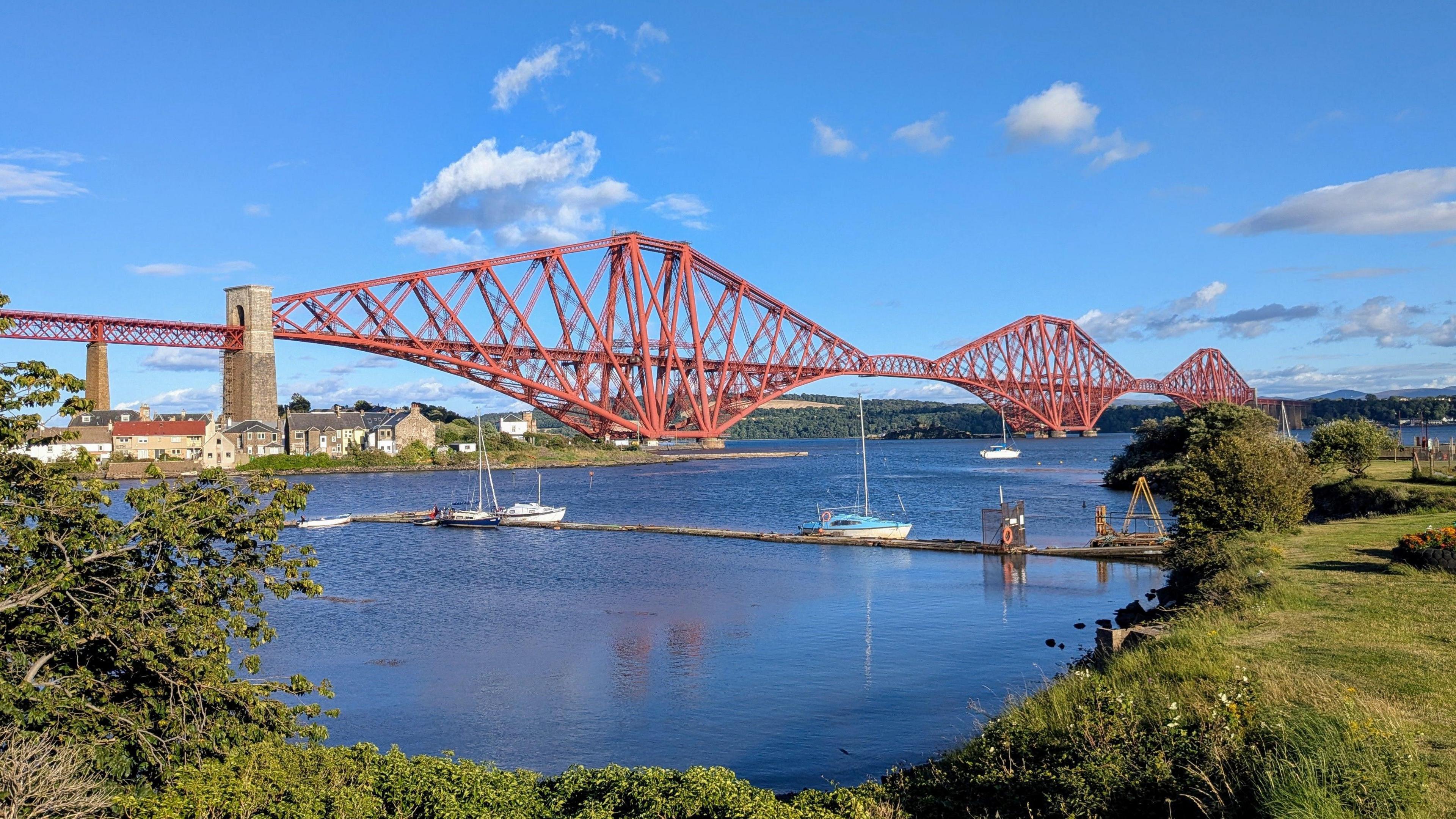 The Forth Rail Bridge is red and stretches across the length of the image. The water below is a dark-blue color, reflecting the sky and the bridge. Small boats are moored in the water near the shore. The shoreline has greenery, including bushes and trees, along the edge of the water, and a grassy area beyond the water's edge.