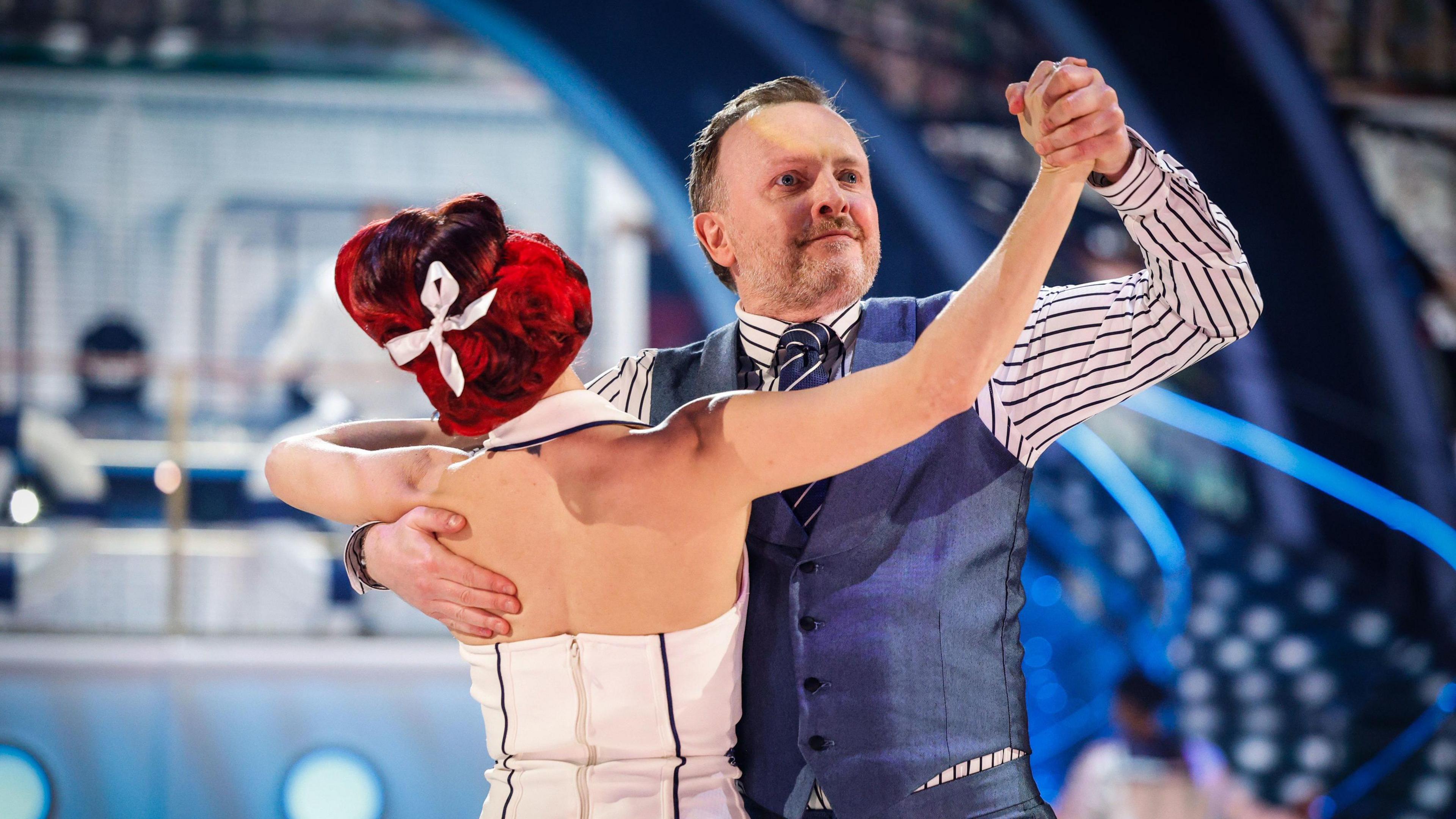 Chris McCausland and his partner Dianne Buswell perform on Strictly Come Dancing. Chris is wearing a blue waistcoat, white striped shirt and a contrasting tie. Dianne has her back to the camera but her red hair has a white ribbon in it. The back of her white and black dress is also visible.