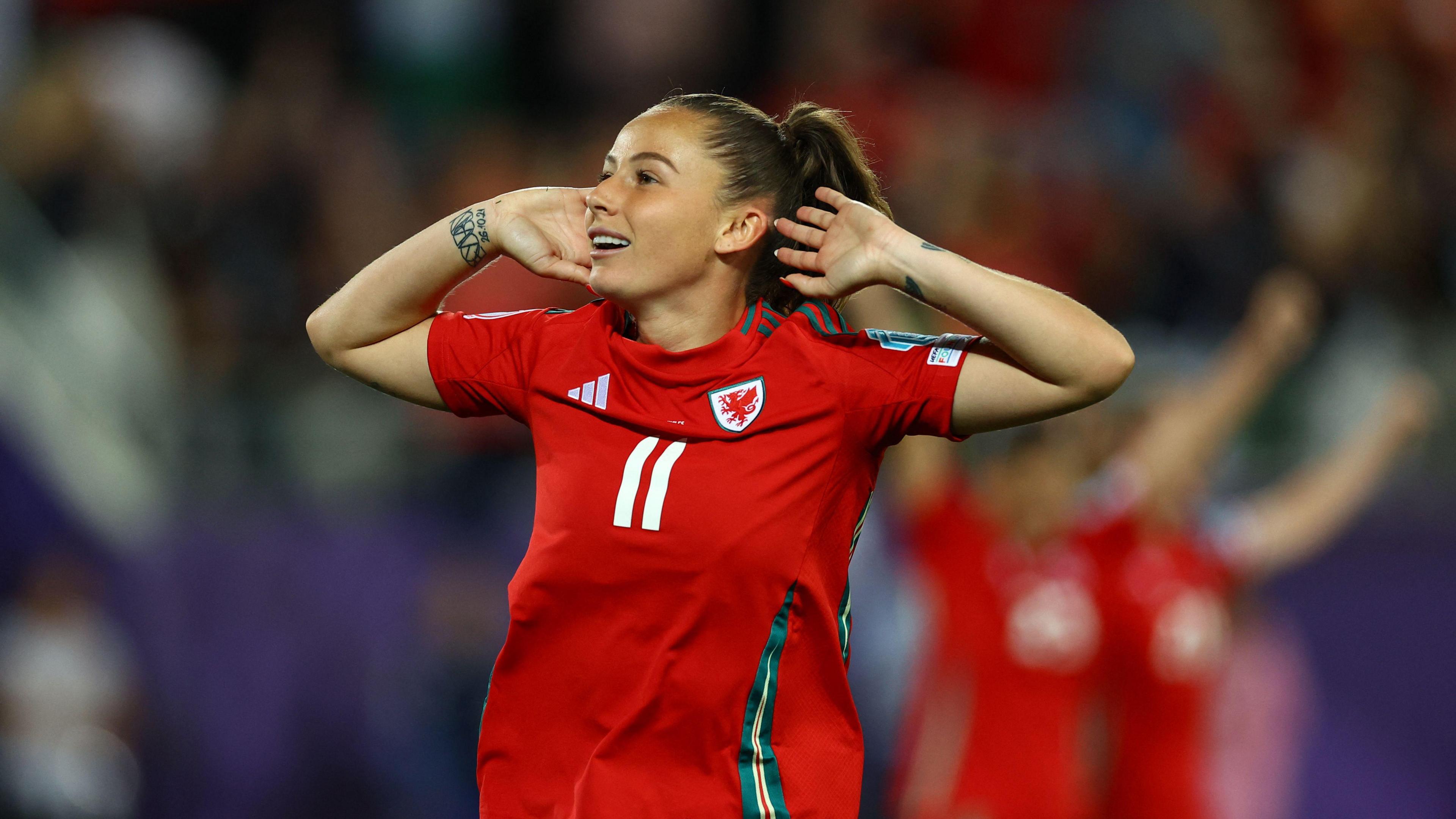 Wales' Hannah Cain celebrates scoring a goal. She holds her hands up to her ears to cup them. She is wearing a red shirt with dark green accents and the number 11 is in white on her chesst
