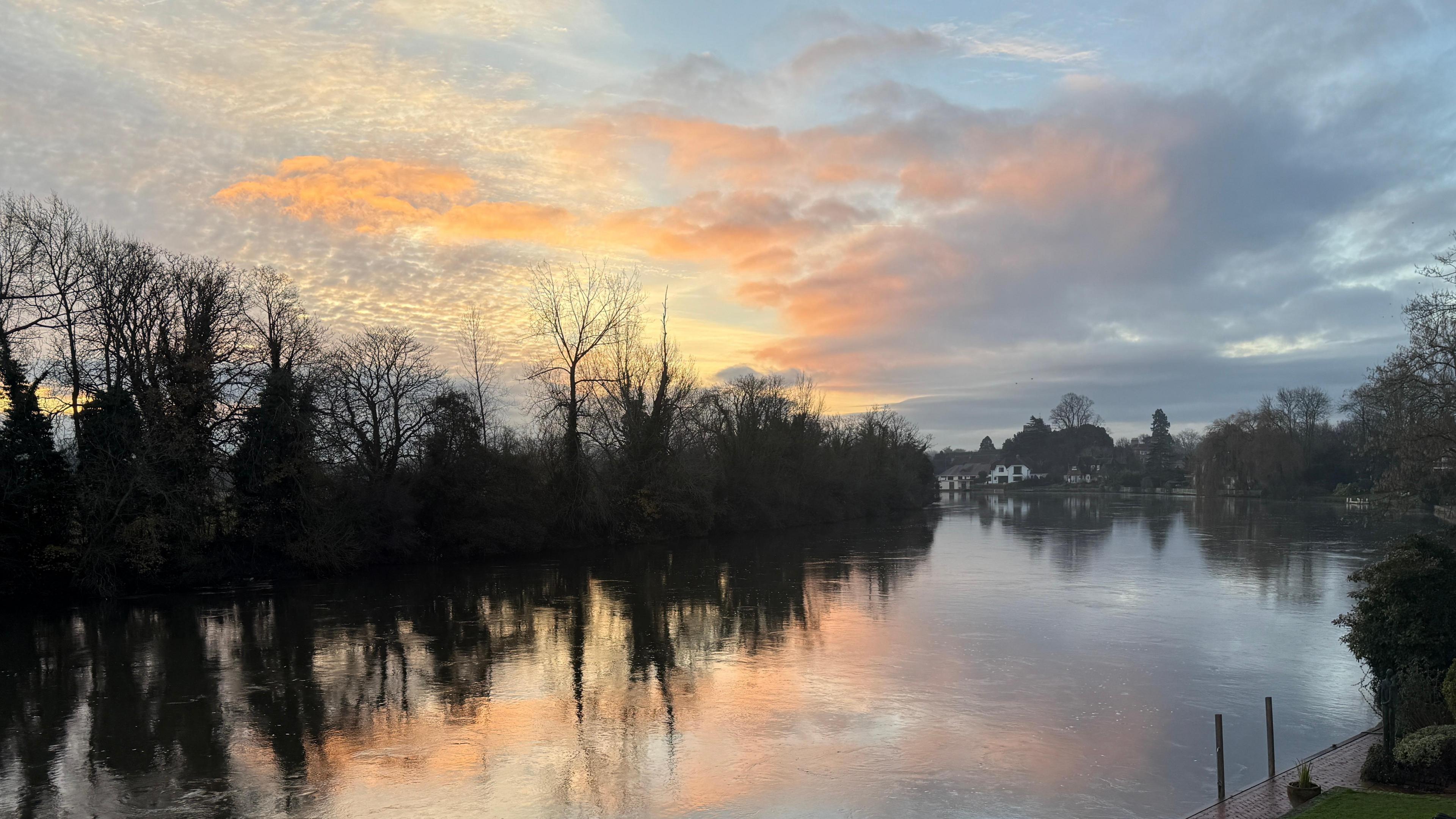 A river at sunrise with a glowing sky overhead. The pink clouds and sun are reflected in the water. On the far bank are trees with bare branches in silhouette against the sky. In the distance you can see white houses on the river bank.  