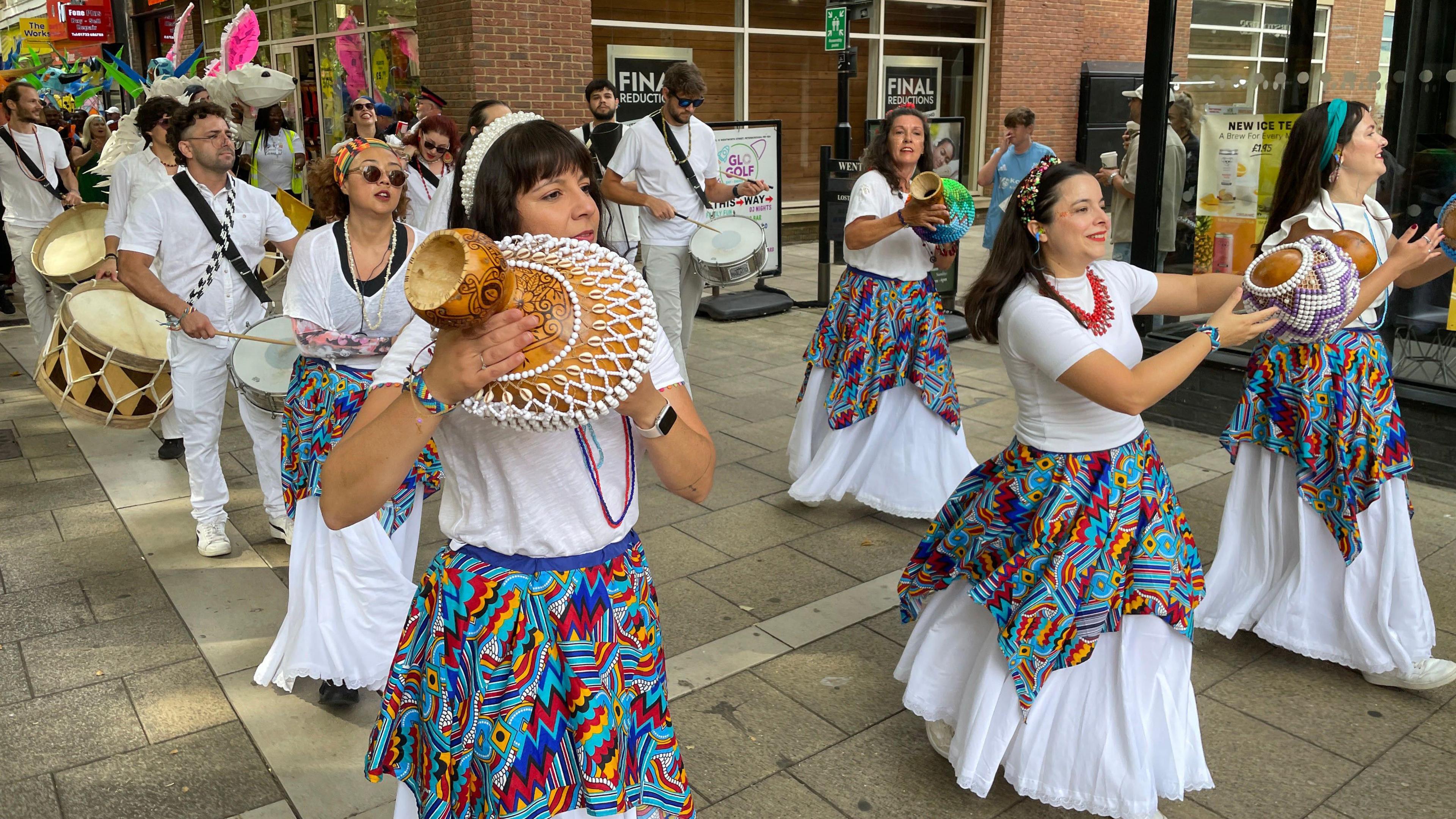 A group of people in colourful costumes, in white and and blues, reds and greens, taking part in a carnival parade in a city centre. They are holding musical instruments. 