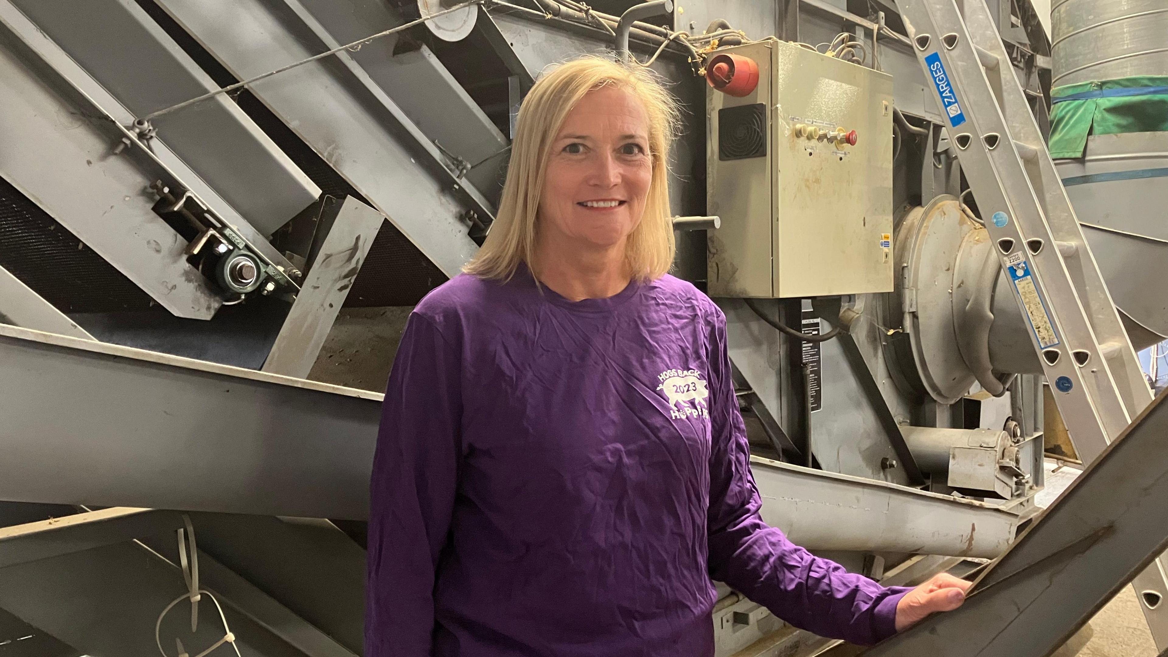 A picture of volunteer Michelle Mackie at the Hogsback Brewery in Tongham, Surrey. Michelle, who has blonde hair and is wearing a purple top, is stood in front of machinery.