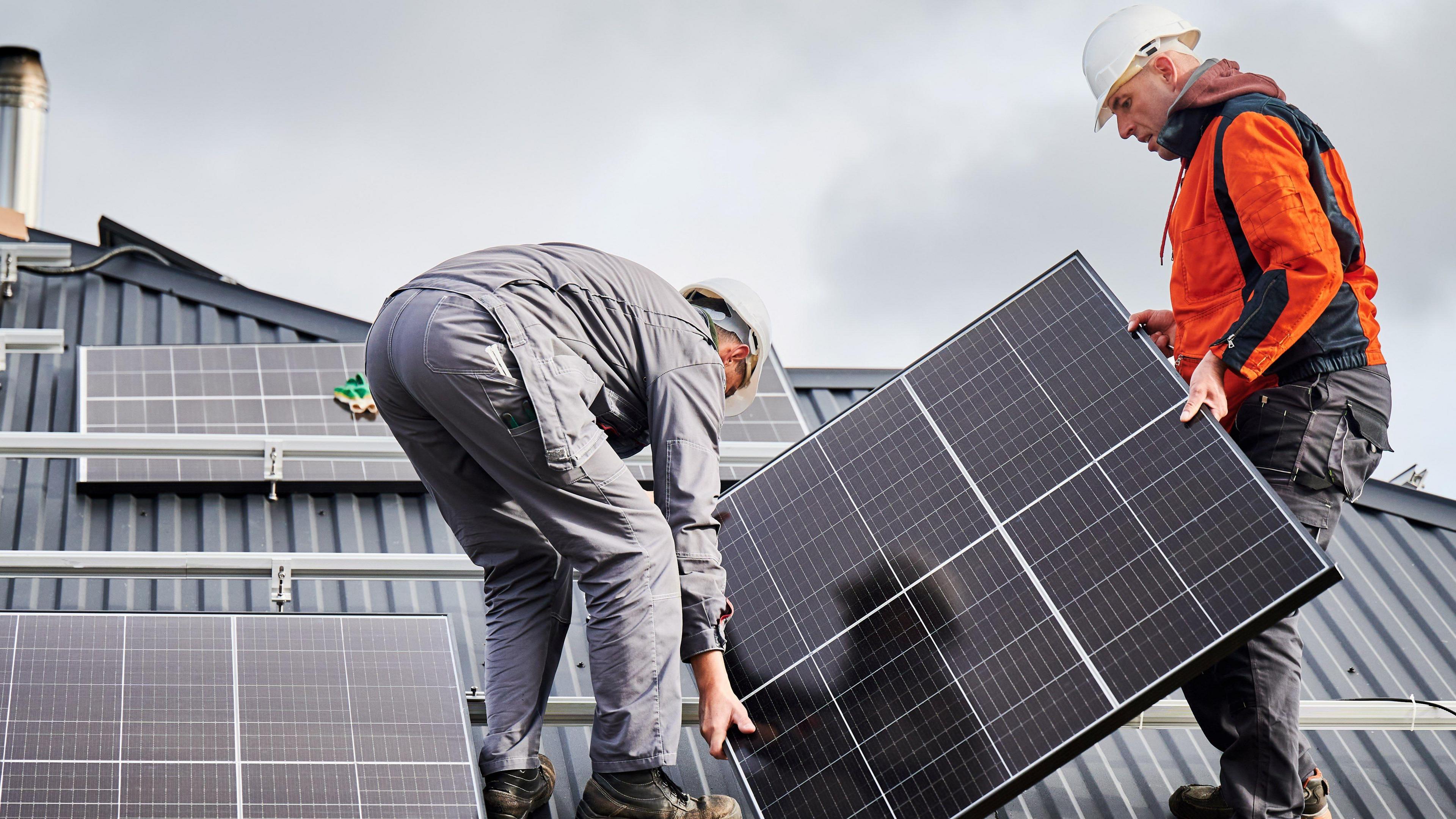 Two workmen standing on a rood. they're installing solar panels. They're wearing hard-hats and boots.