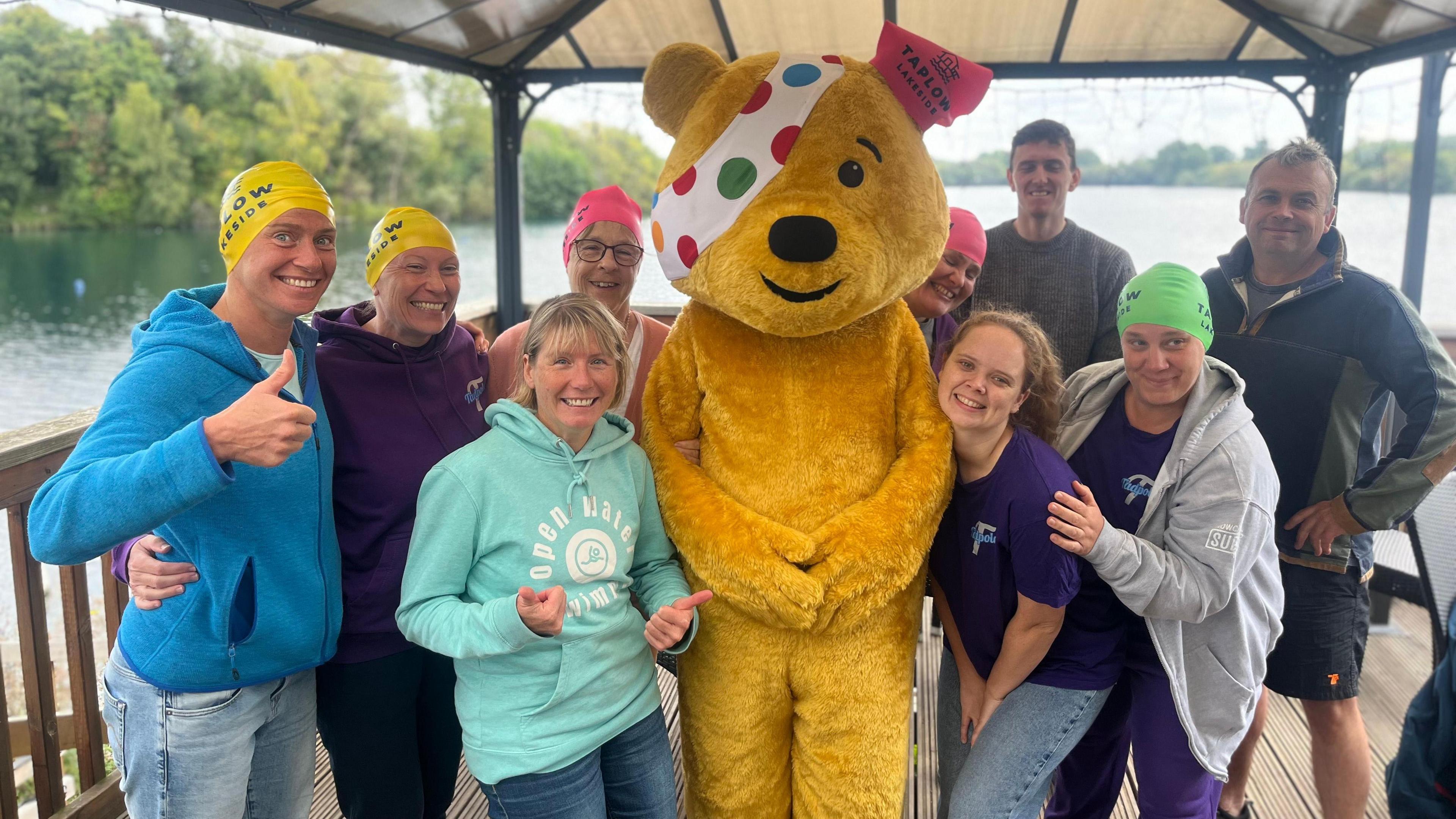 A group of people, some wearing swimming hats, smiling and posing for a photo with someone in a Pudsey Bear costume. There's a lake in the background.