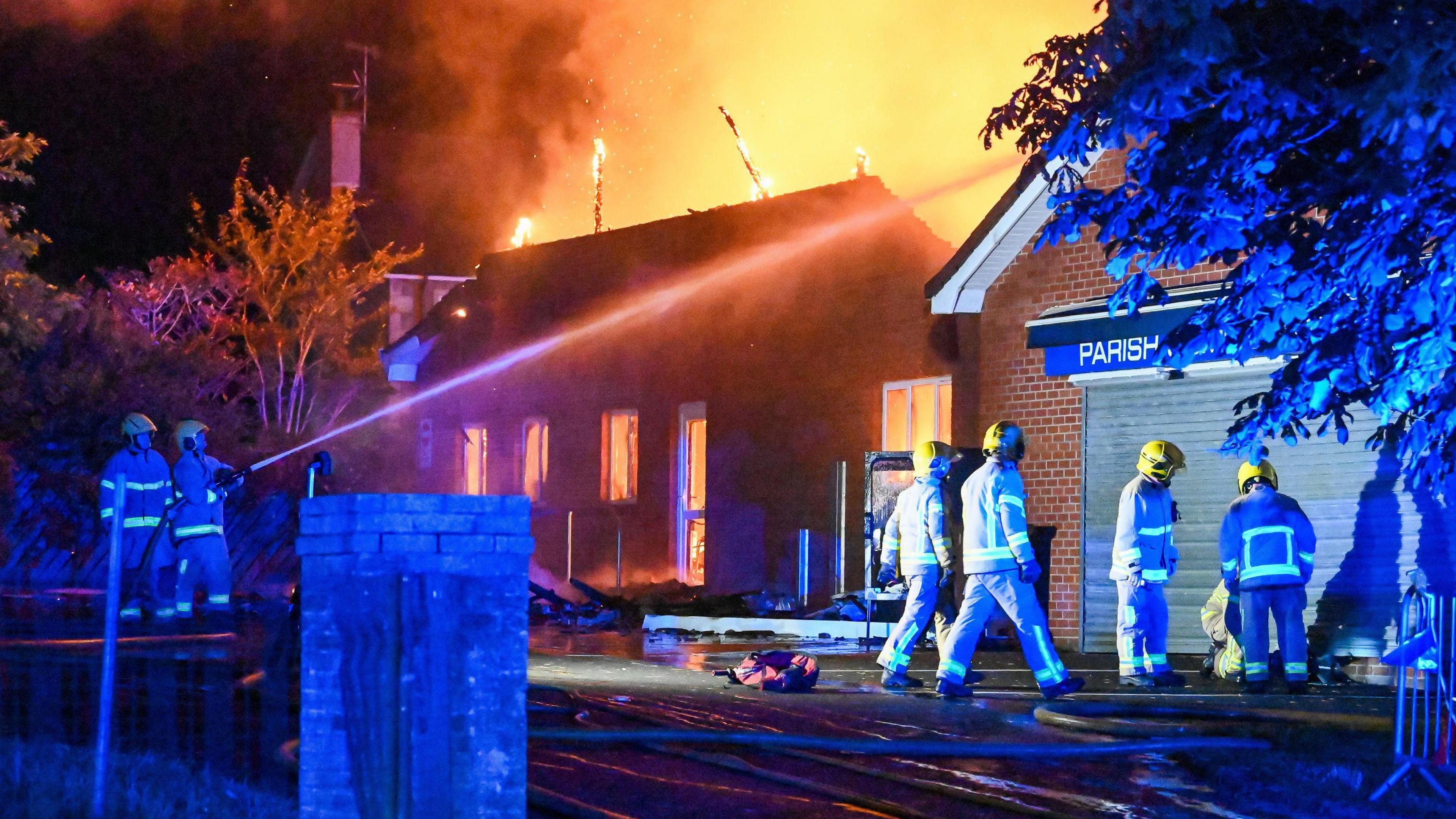 Firefighters fighting a blaze at the church in Greenisland last weekend. One firefighter is trying to extinguish the fire with a hose. The windows are red with the fire and smoke is in the air.