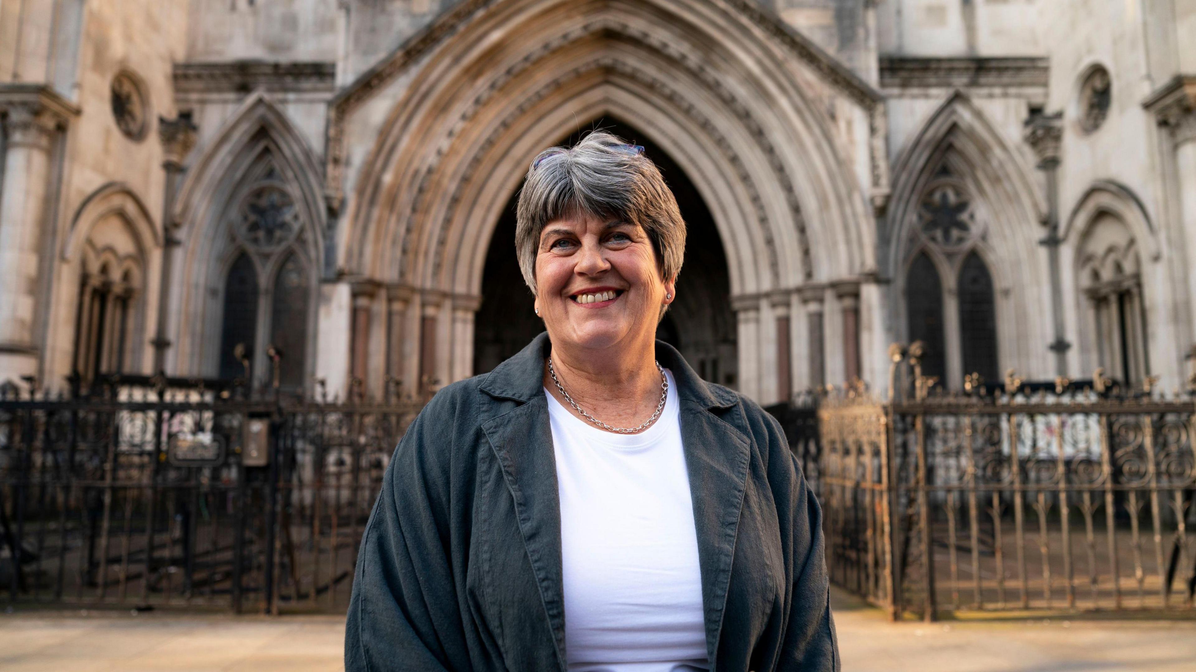 A woman stands outside an ornate carved stone building. She has short grey hair and is wearing a dark green linen jacket and white tee-shirt. 