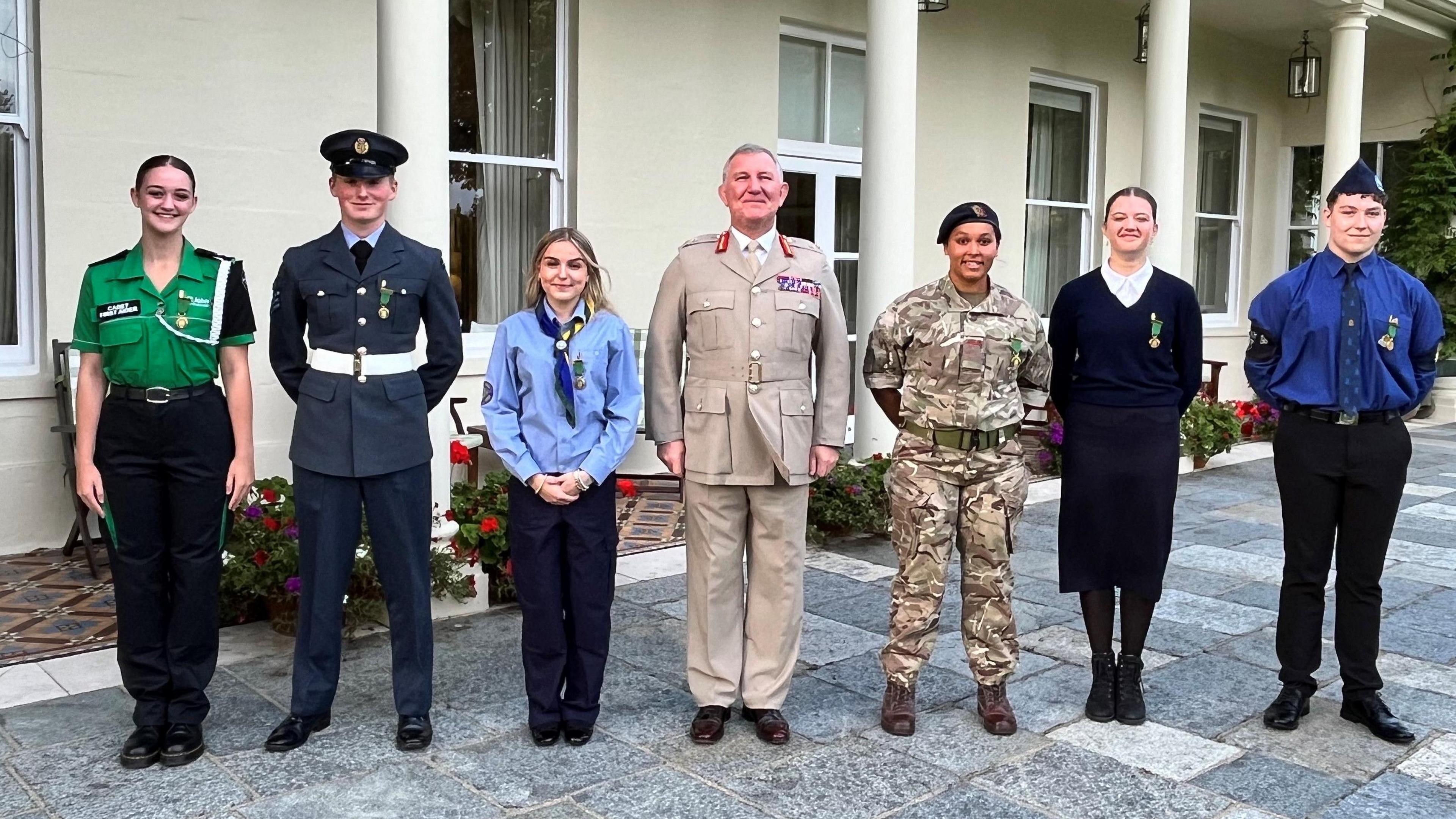 The six cadets, each wearing their different uniforms, stand outside a pillared building looking at the camera with the Lieutenant Governor in the centre of the line-up.