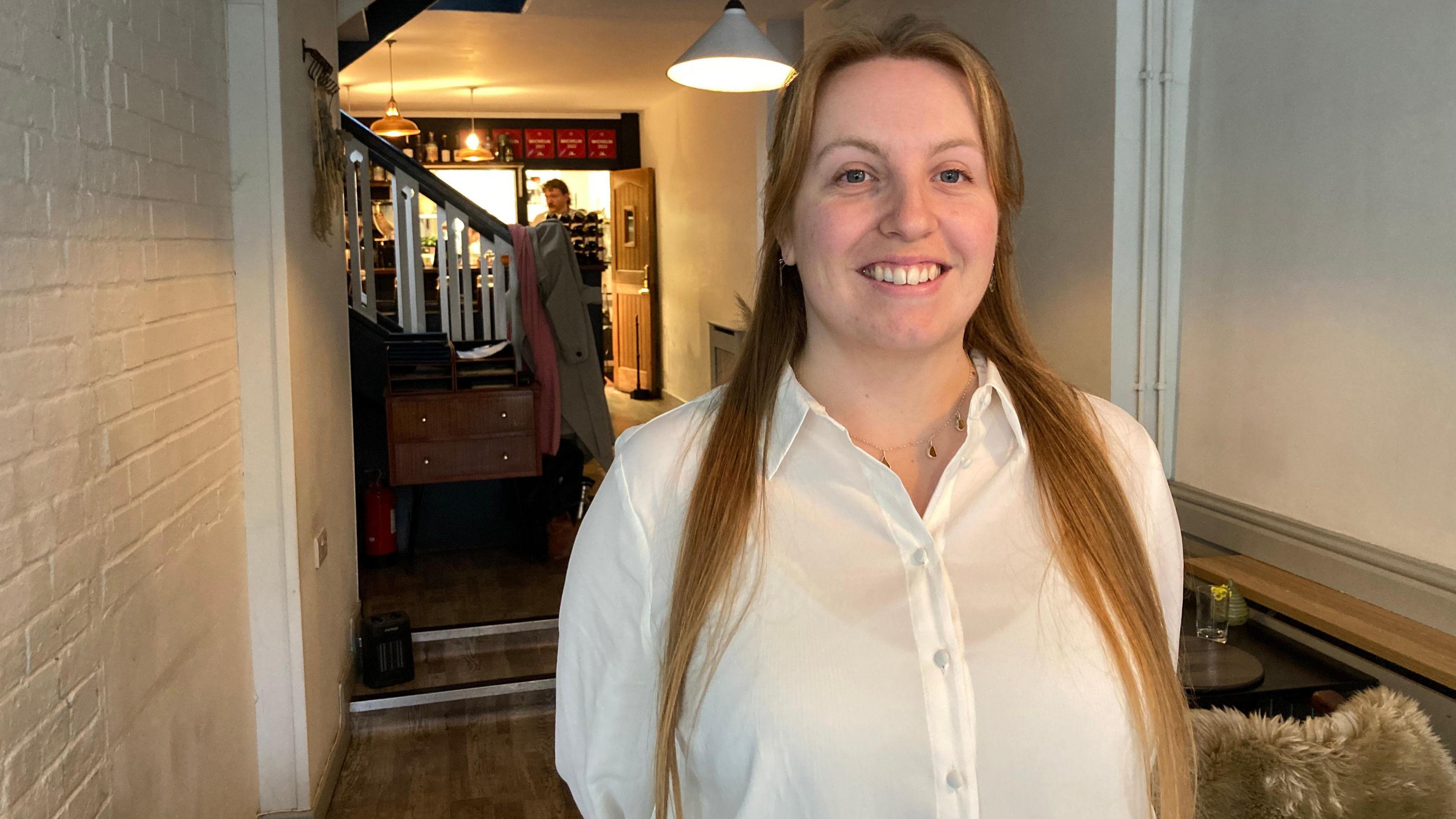 A woman with long blonde hair wearing a white shirt is smiling with her hands behind her back. She is standing in a restaurant. 