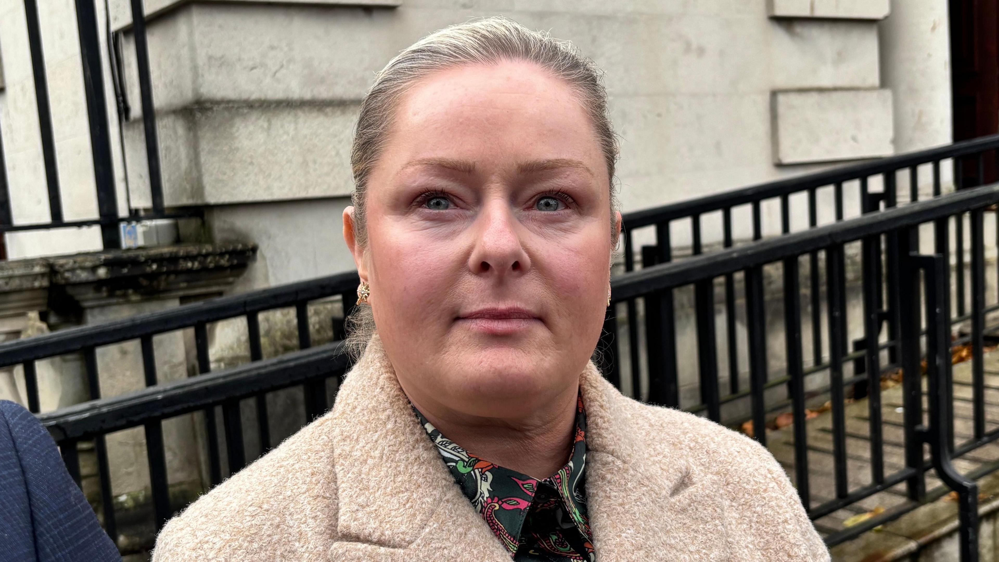 Ann McClure looking at the camera. She is wearing a beige coat and shirt that is green with pink, white and orange pattern. Behind her there is a cream coloured wall with black railings in front of it.
