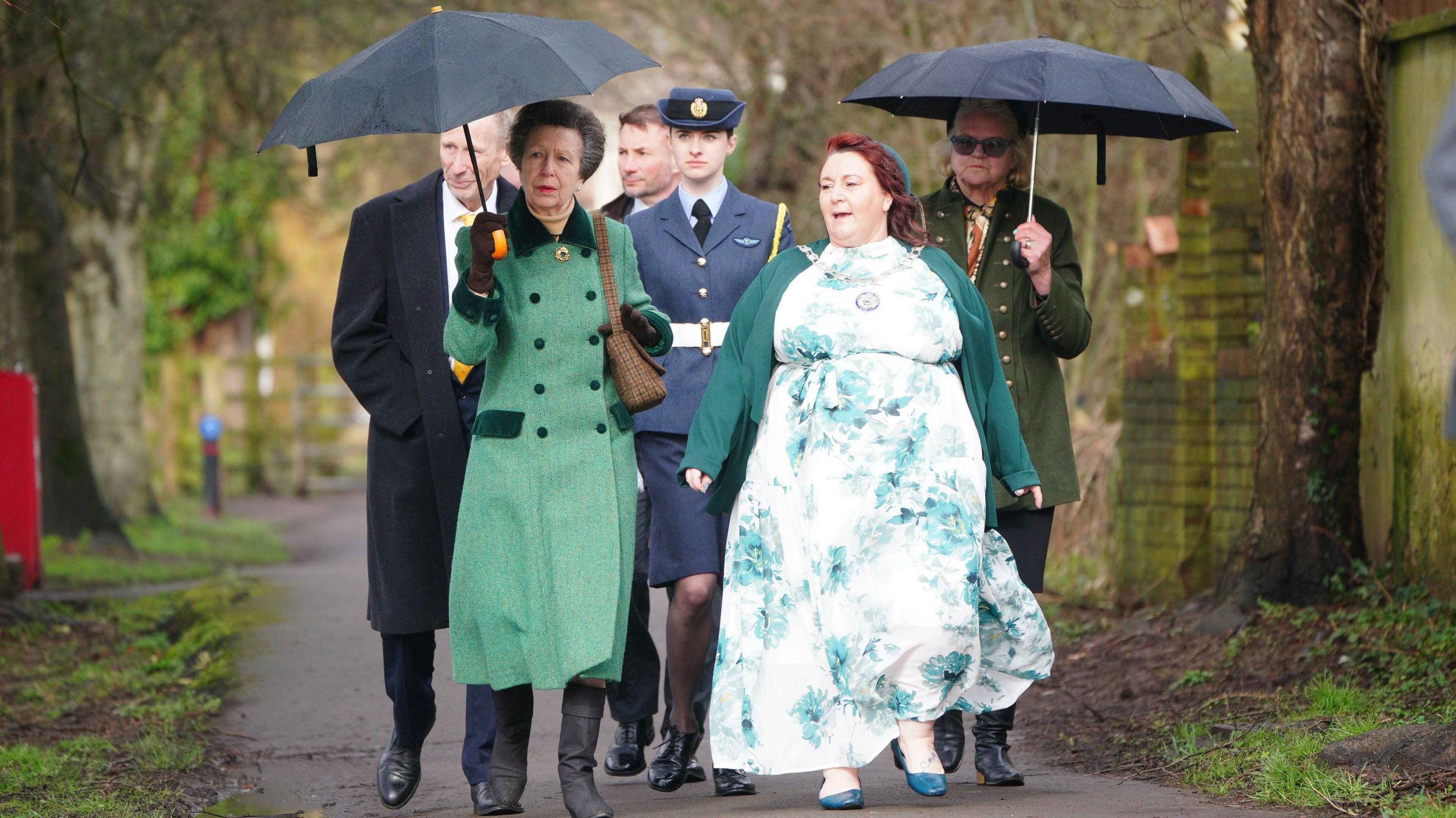 Princess Anne, holding a black umbrella and wearing a long light green coat with dark green details, walks with a group of people along a path through a park. 