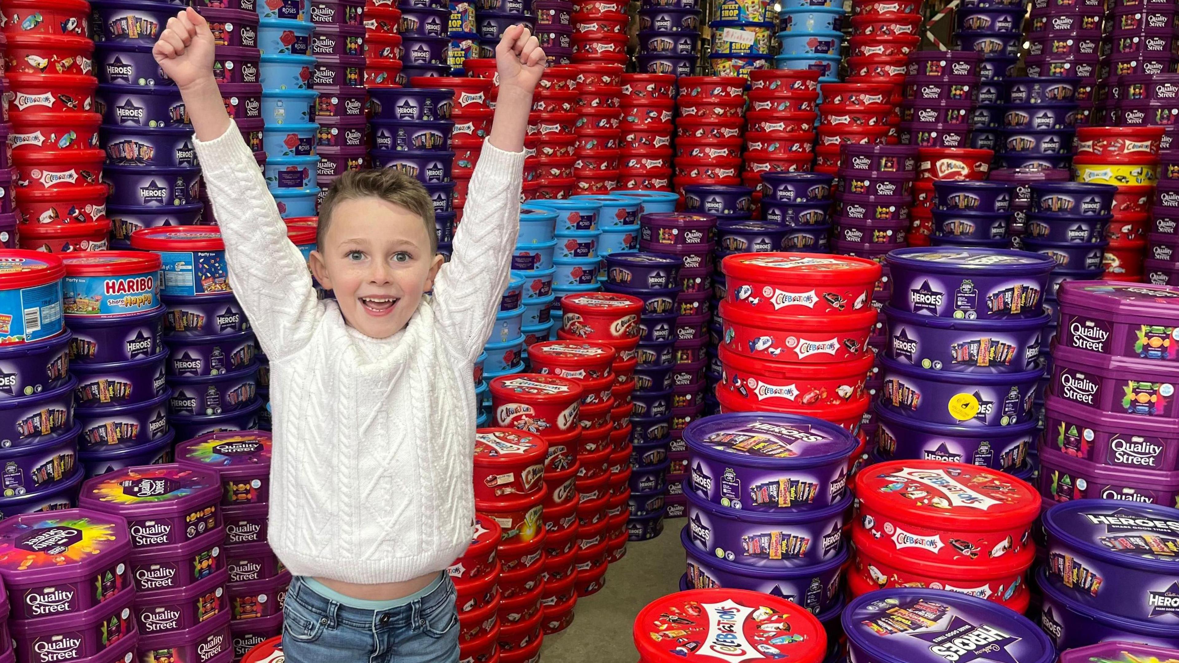 A boy in a white jumper smiles surrounded by hundreds of plastic tubs which contained sweets and chocolate