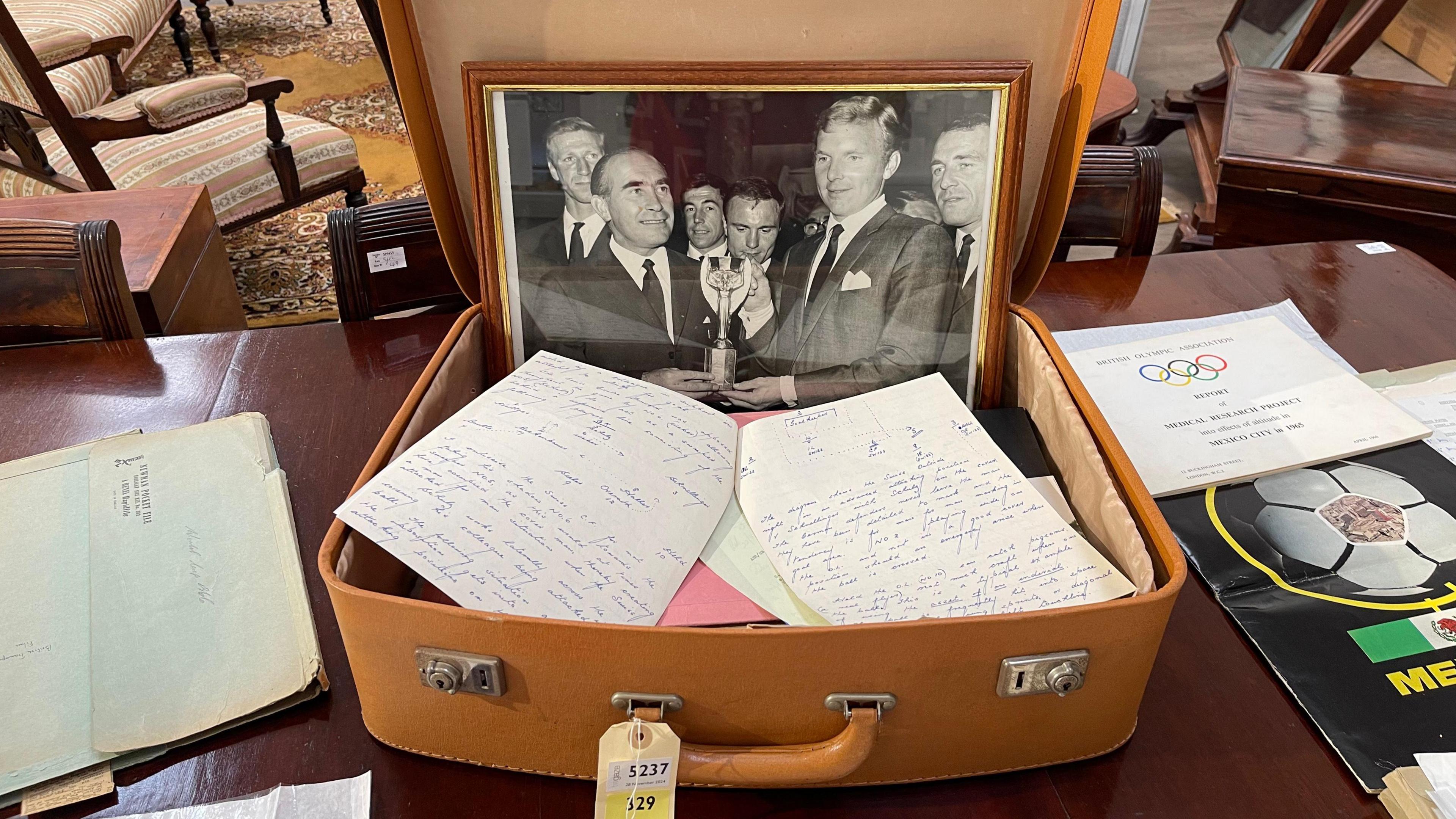 A brown suitcase with a black and white photograph in a frame, on a brown wooden table with other pieces of papers with writing on it and programmes.