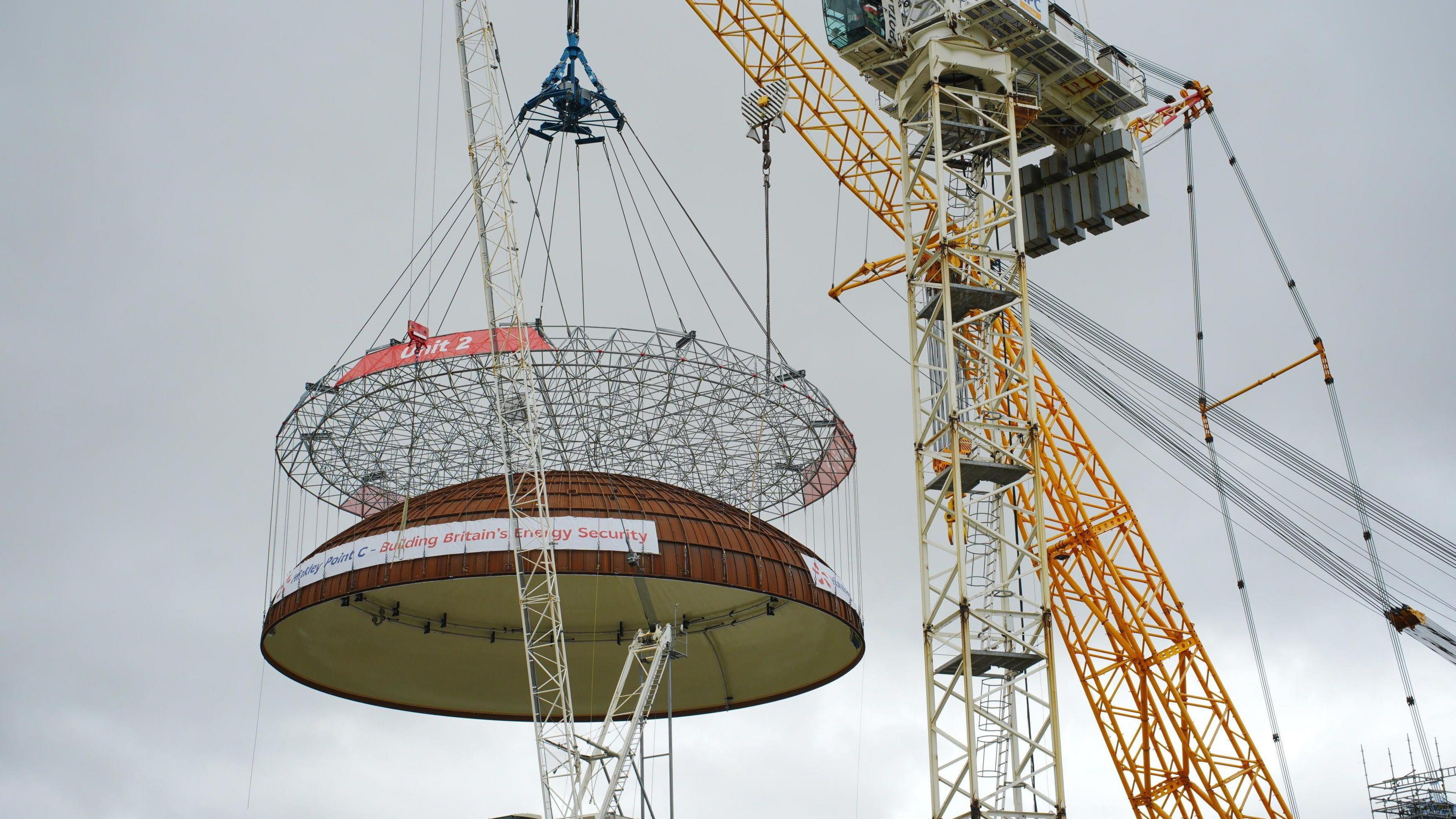 A close up image showing the reactor dome being slowly lowered onto the building below using a large yellow crane. The sky behind it is dark and cloudy.