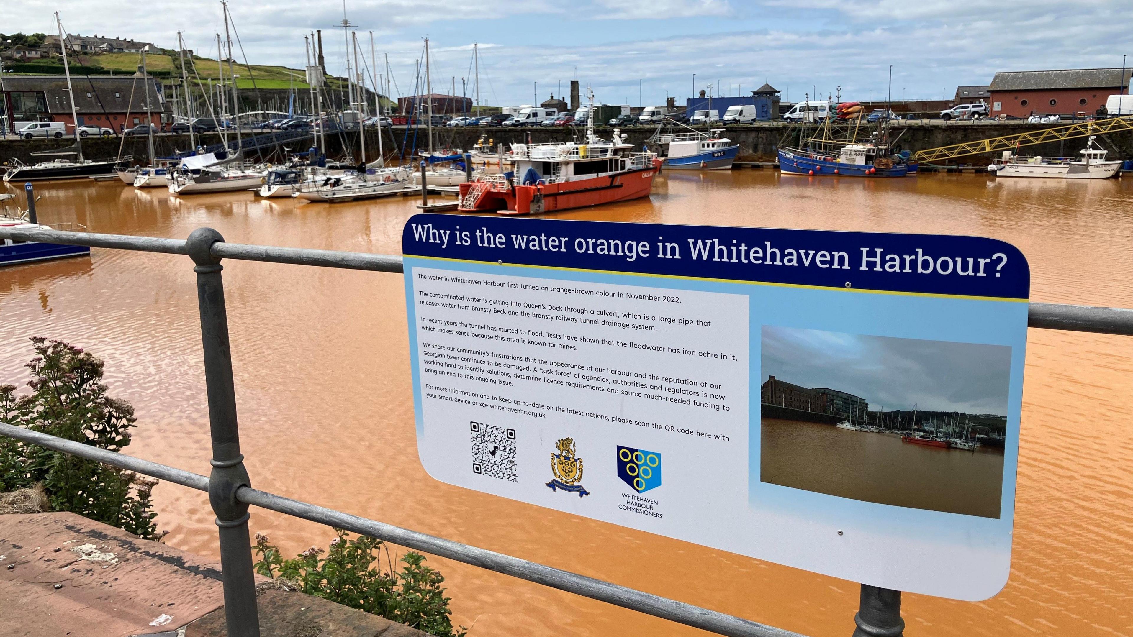 An image of a sign on barriers at Whitehaven Harbour explaining why the water is orange. Behind the board is the marina, with boats moored and orange water.