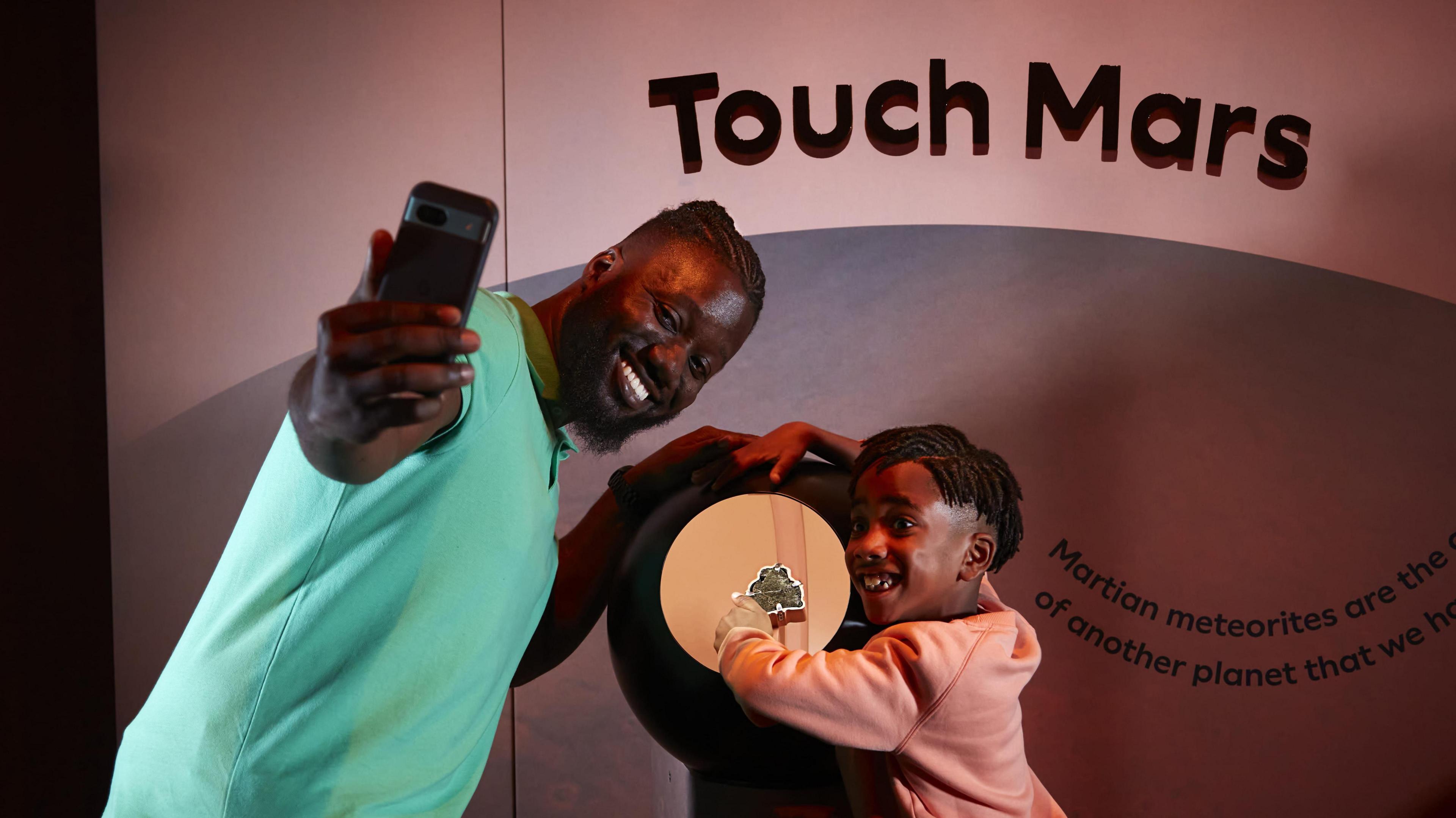 A man and a child pose for a selfie with an iPhone in front of a meteorite from Mars.