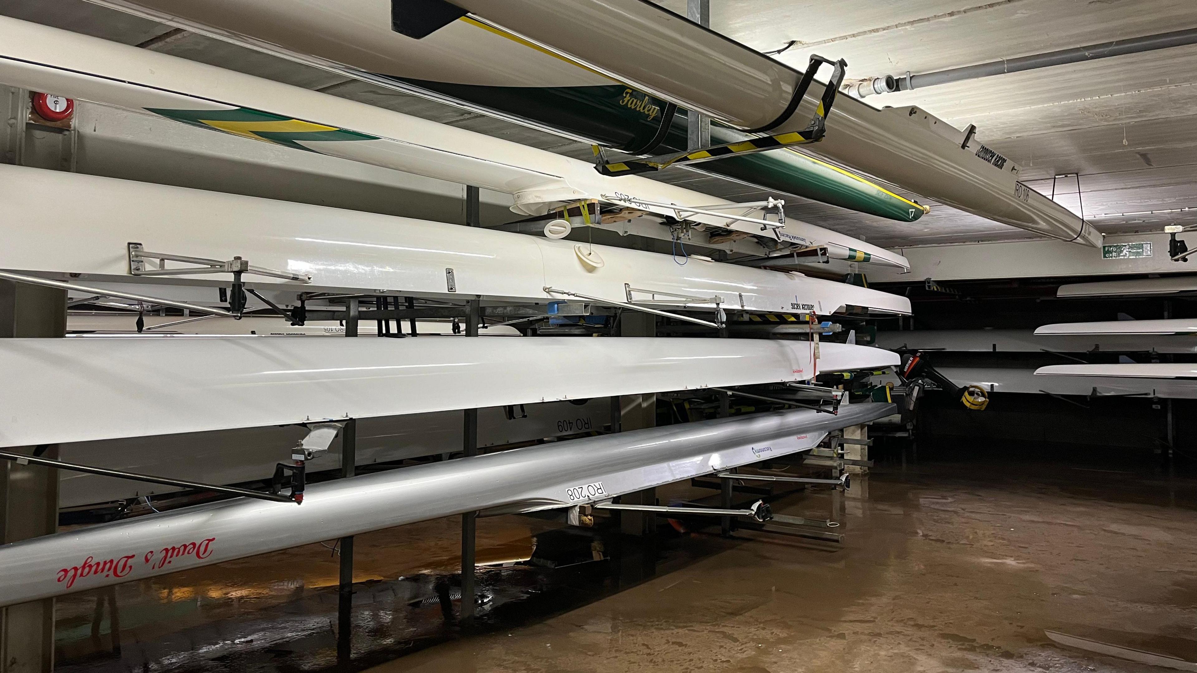 Long white rowing boats are stacked inside a boathouse. There is brown flood water underneath the stack.