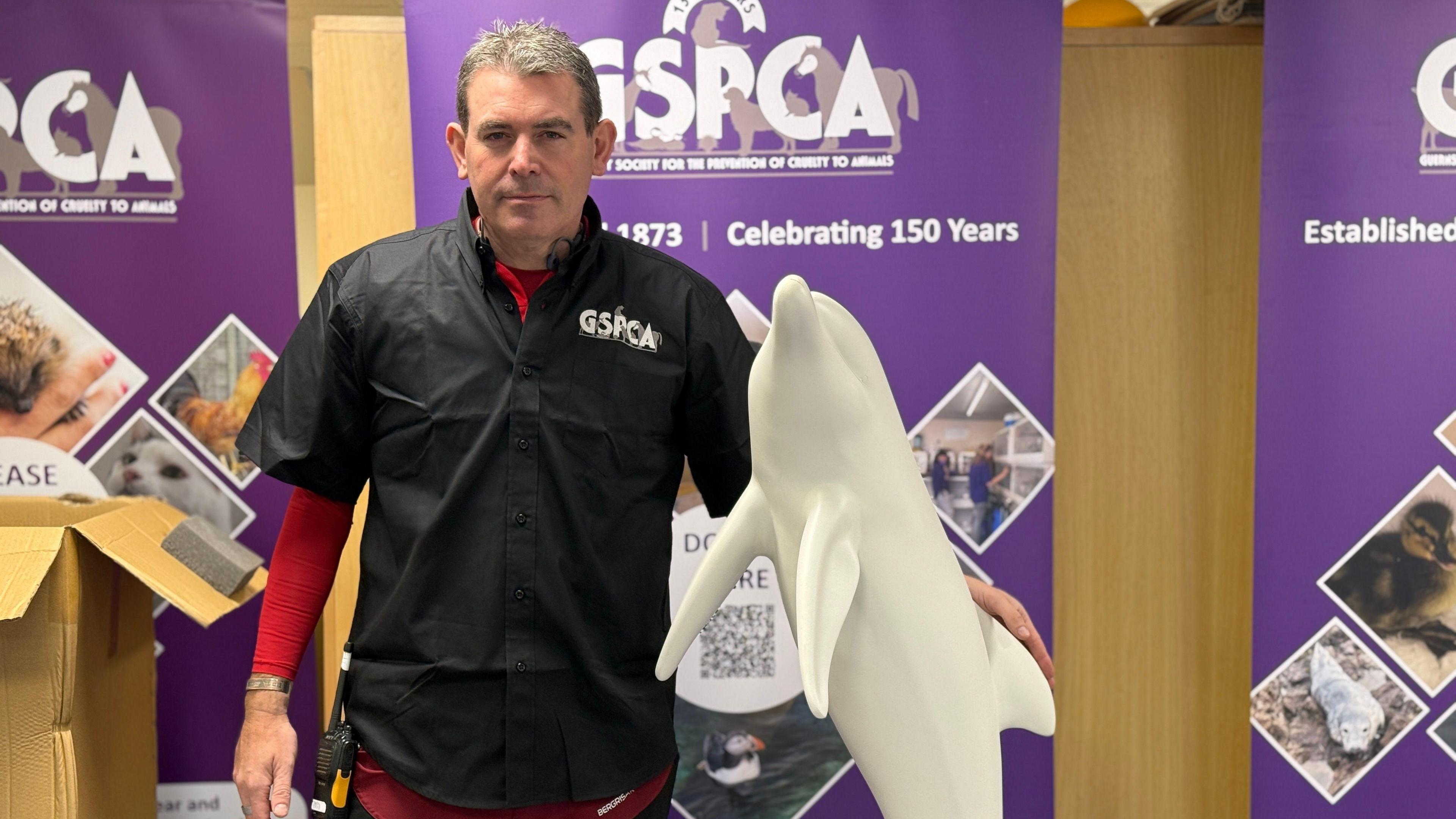 GSPCA manager Steve Byrne, who is a middle-aged man with grey hair and wearing a black short-sleeved shirt with the GSPCA logo with a red under layer. He is standing next to a white sculpture shaped like a dolphin.