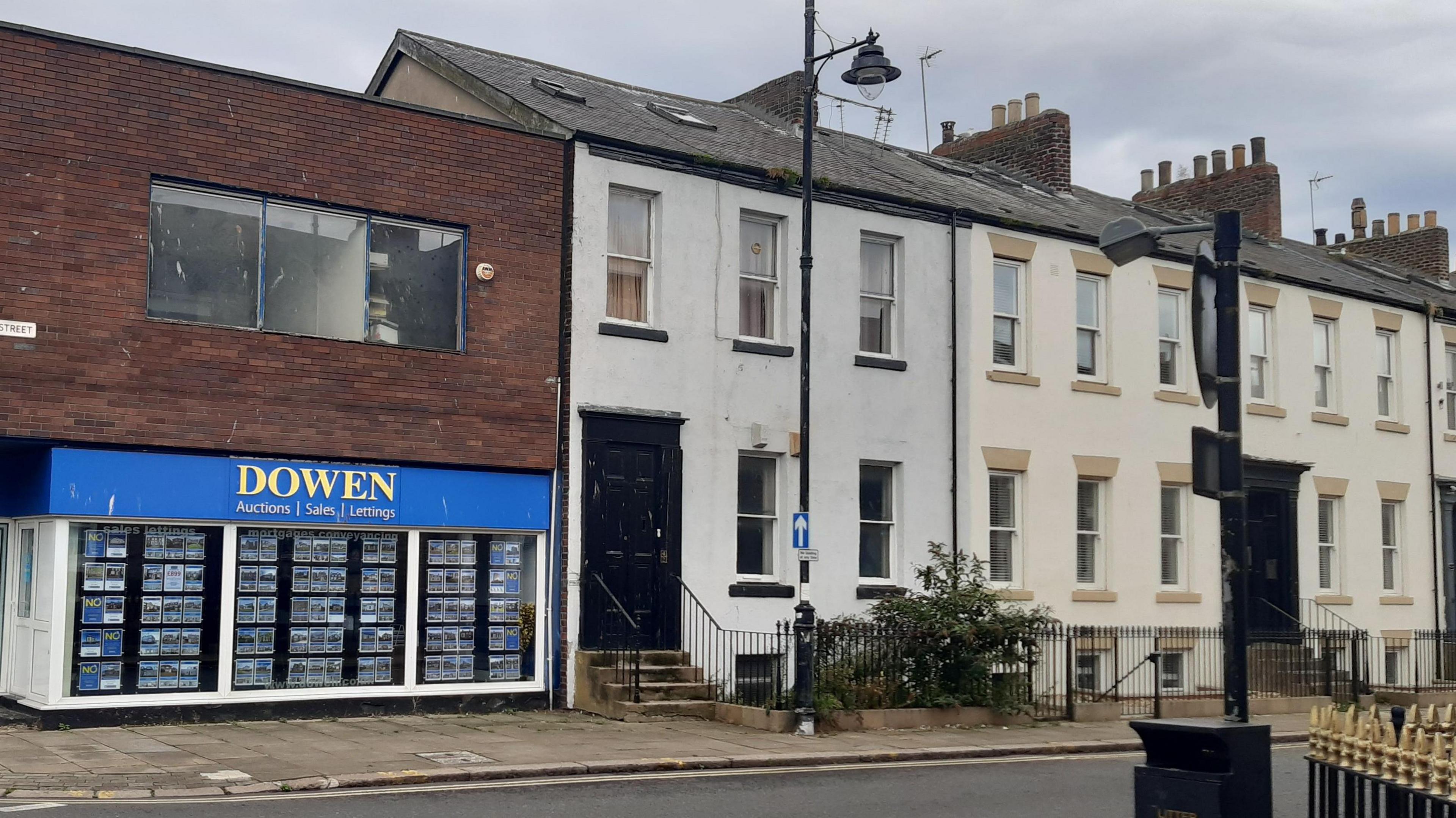 42 Frederick Street, Sunderland. A two-storey grey-white building with large, black front door and black detail around the windows, of which there are two on the ground floor and three on the first. Two skylights are visible on the roof. To its left is a modern, flat-roofed estate agent shop with a blue and yellow sign and a rectangular, plain window set in the brick-fronted upper floor.