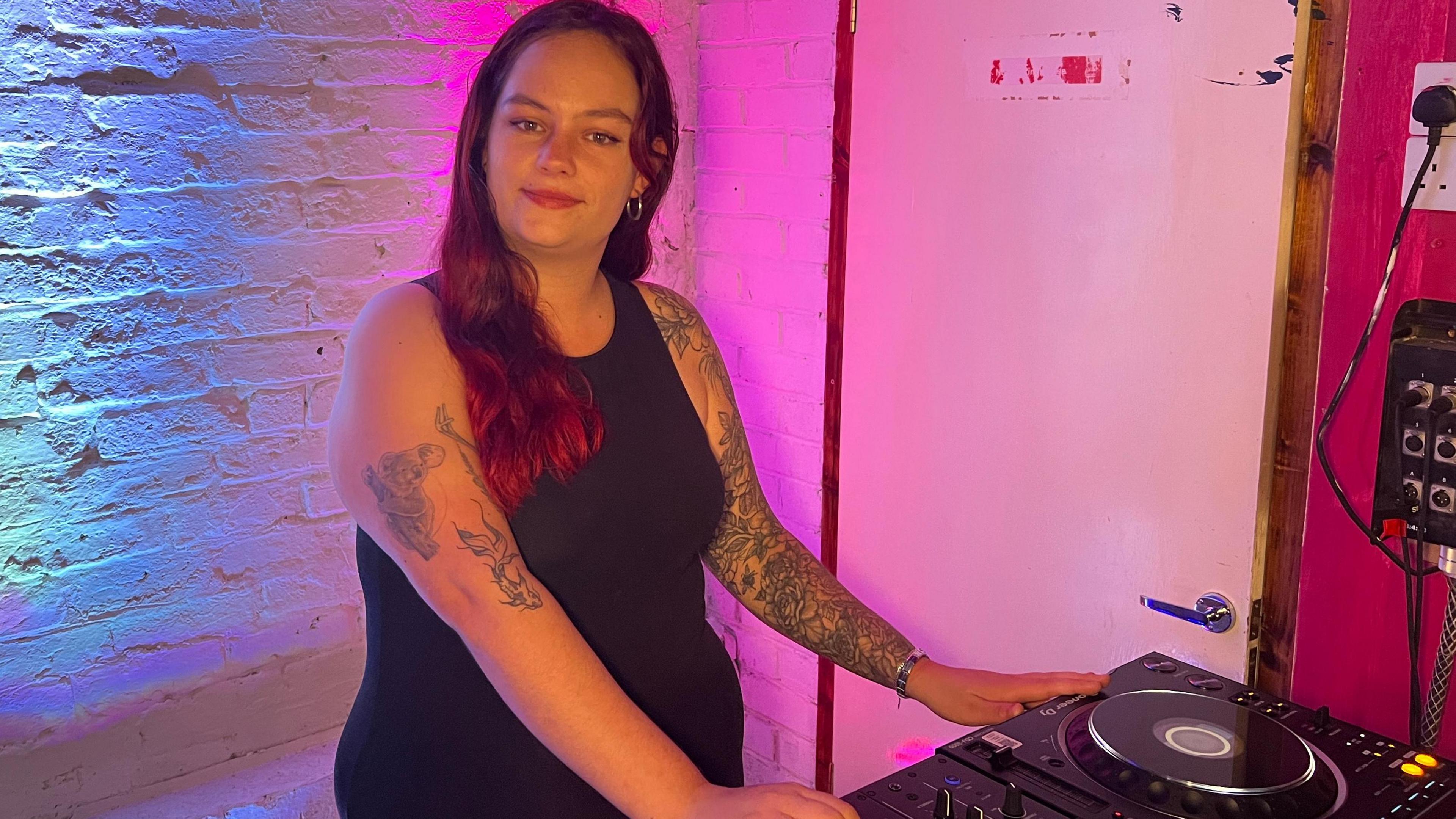 A white woman with long, red hair, stood over a set of DJ decks. She has a full sleeve tattoo on her right arm, and a tattoo of a fish and a koala on her upper left arm. She is smiling and is wearing a black vest and black trousers.