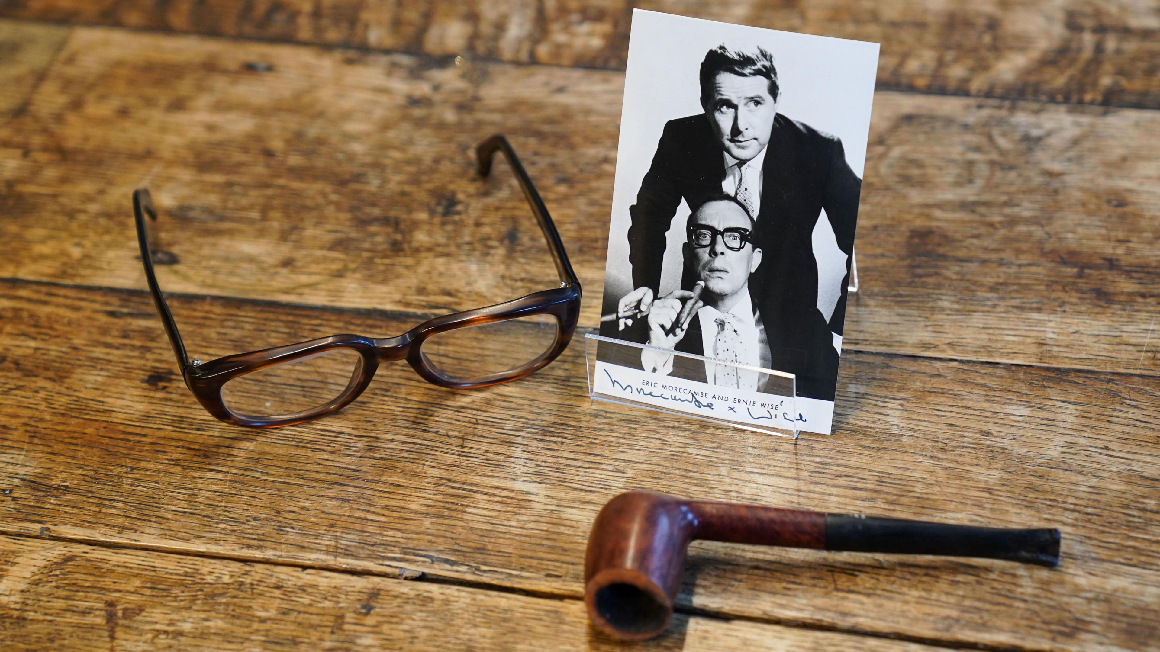 Eric Morecambe's glasses and pipe resting on wooden planks. The glasses are brown imitation tortoiseshell and their arms are open. The pipe is lying on its side and is brown. There is also a black and white photo showing Eric Morecambe with Ernie Wise. 