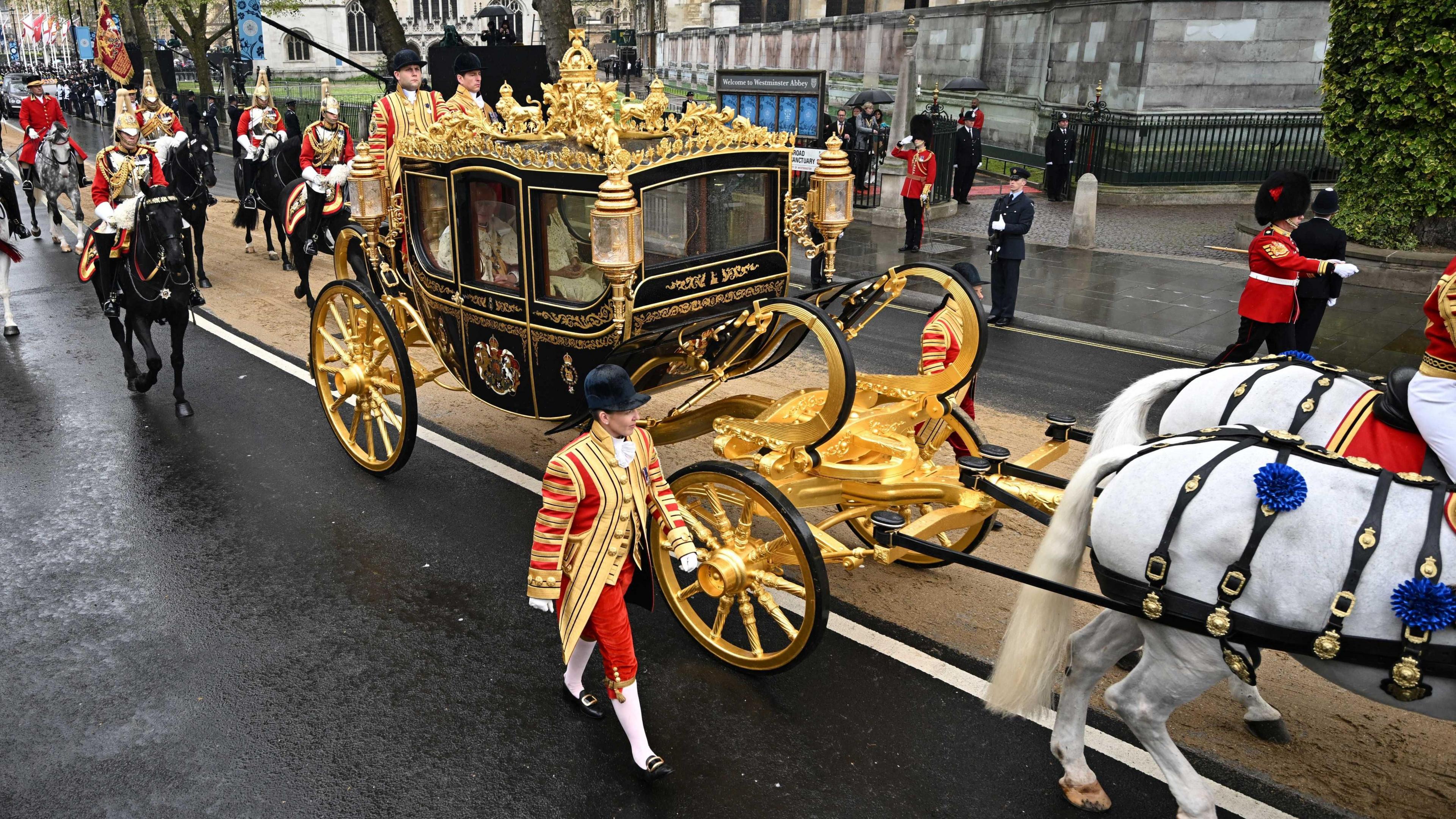 A modern coach being pulled along a London street by white horses. Liveried staff walk beside the coach and there are mounted cavalry in the rear. The King and Queen are visible through the windows.