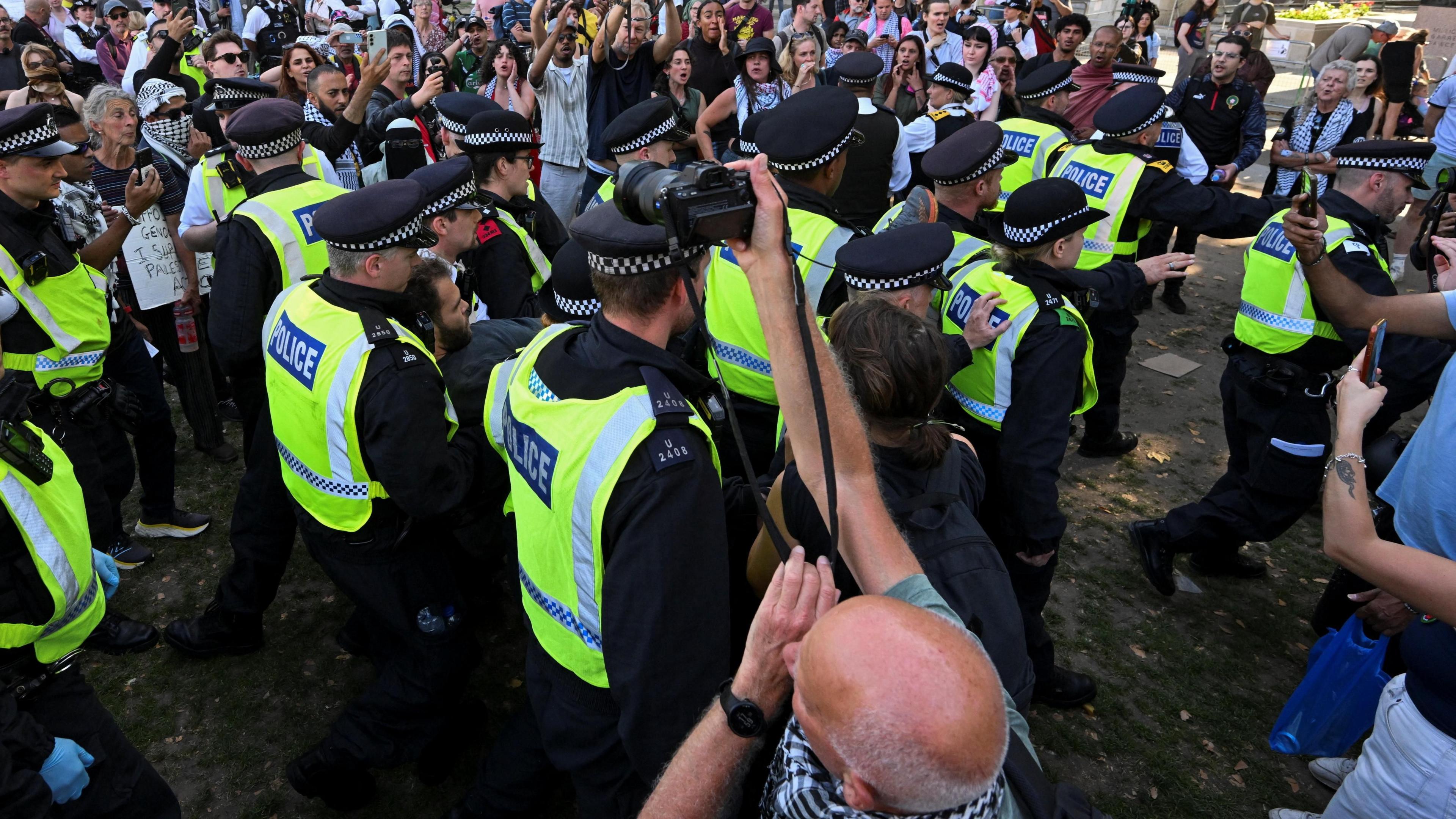 Police officers detain a protester during a rally aimed at challenging the government's proscription of Palestine Action under anti-terrorism laws, in in London, Britain on 9 August. The picture shows a cluster of police in high-vis jackets, holding a man, with a crowd surrounding them. In the foreground, a man takes pictures with a camera.