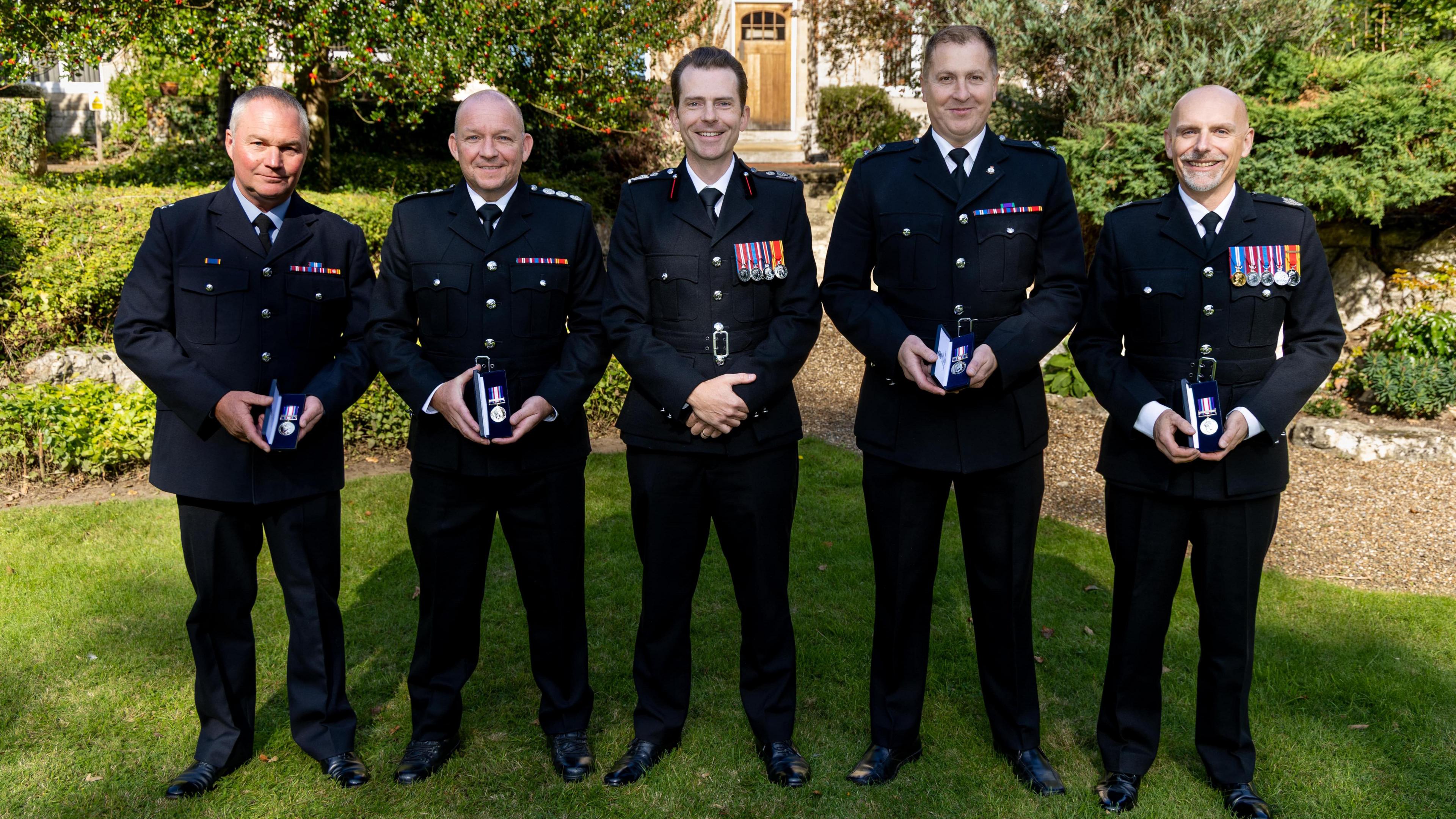 Five men in formal black firefighter uniform suits, stood in the sun in a beautifully manicured garden. All but the man in the centre hold small silver medals in presentation boxes.