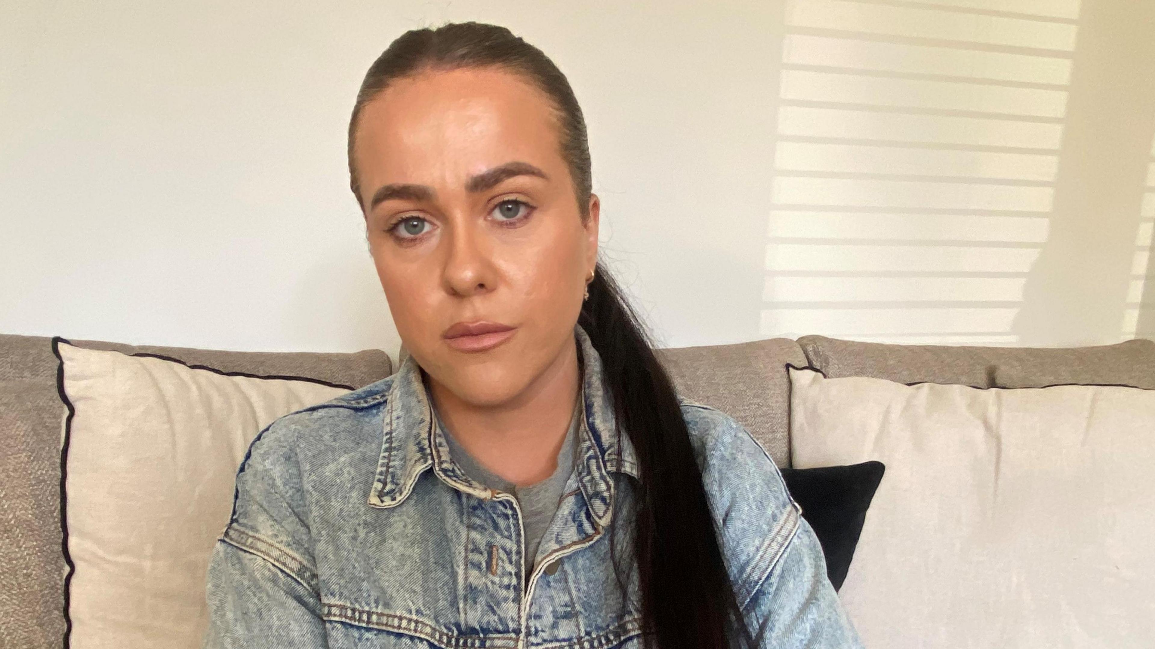 Head and shoulder shot of a young woman. She has long dark hair, tied tightly back in a pony tail which drapes over her left shoulder, and is wearing a denim jacket.  Her expression is serious. She is sitting on a sofa, with the back a light brown linen weave type material and with cream cushions.