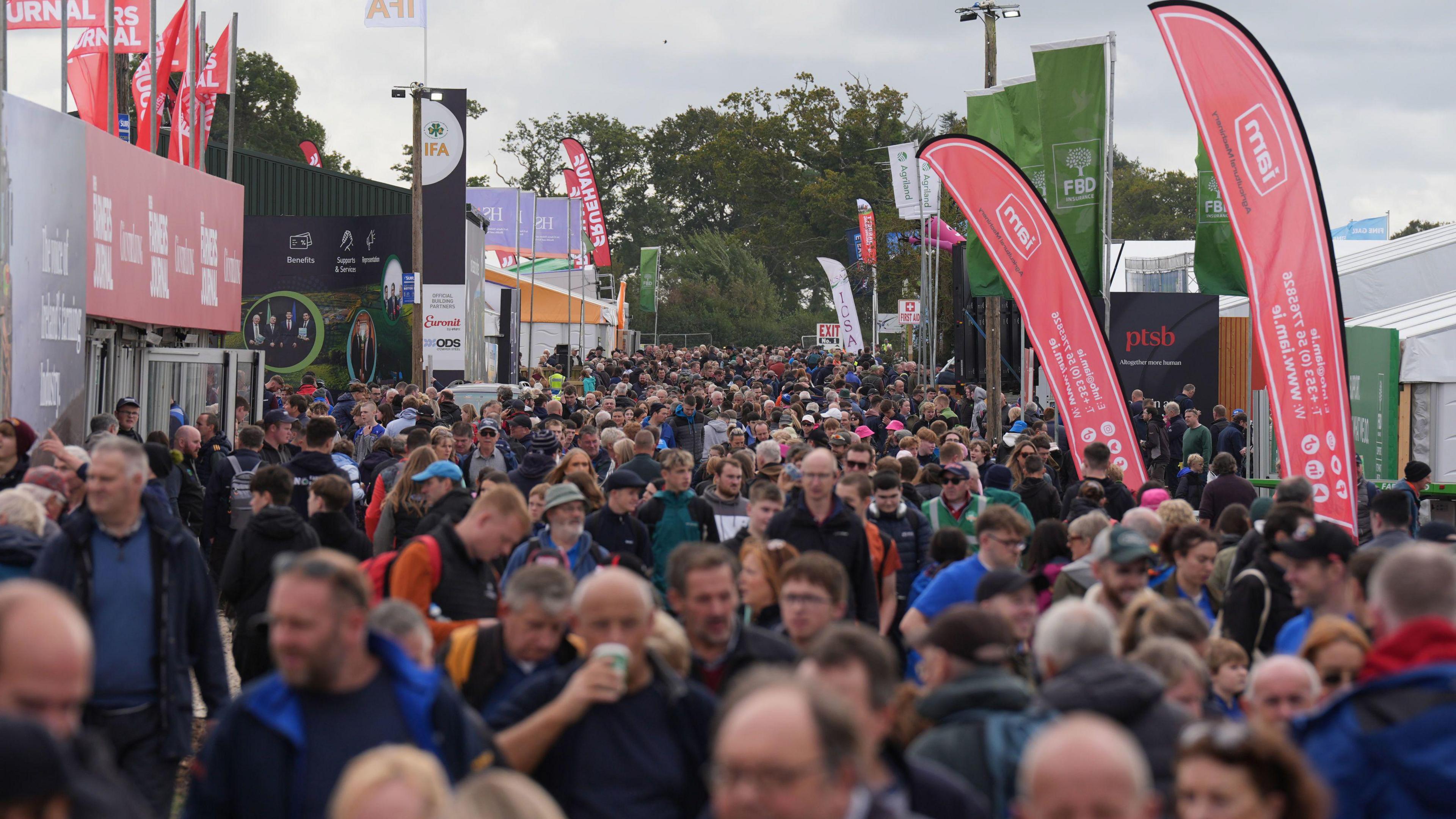 Crowds of people queue with stalls and banners on either side. The sky is grey.