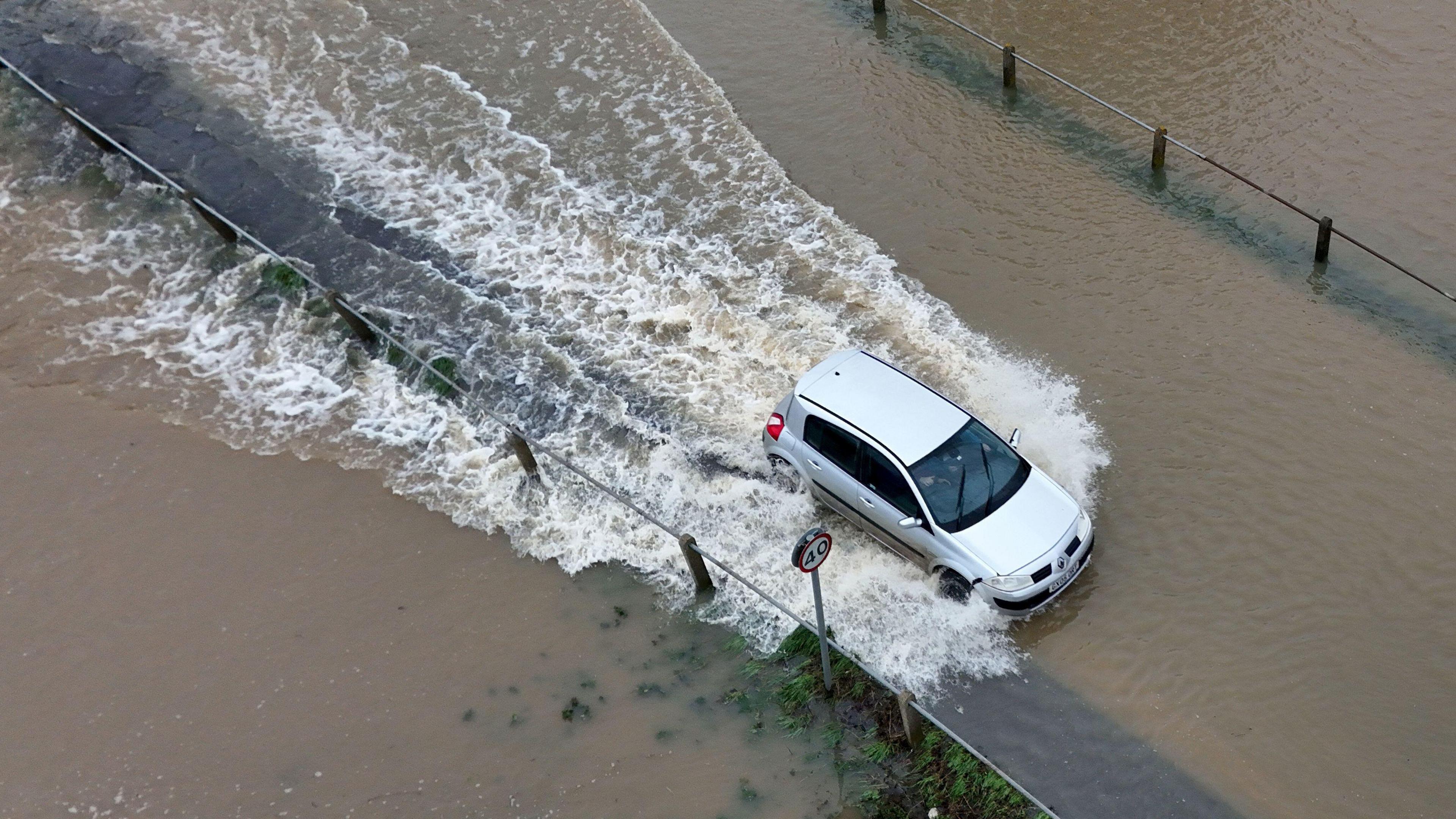 A car driving through a flooded road with water splashing around, partially submerging the road surface, and a 40 mph speed limit sign visible nearby.