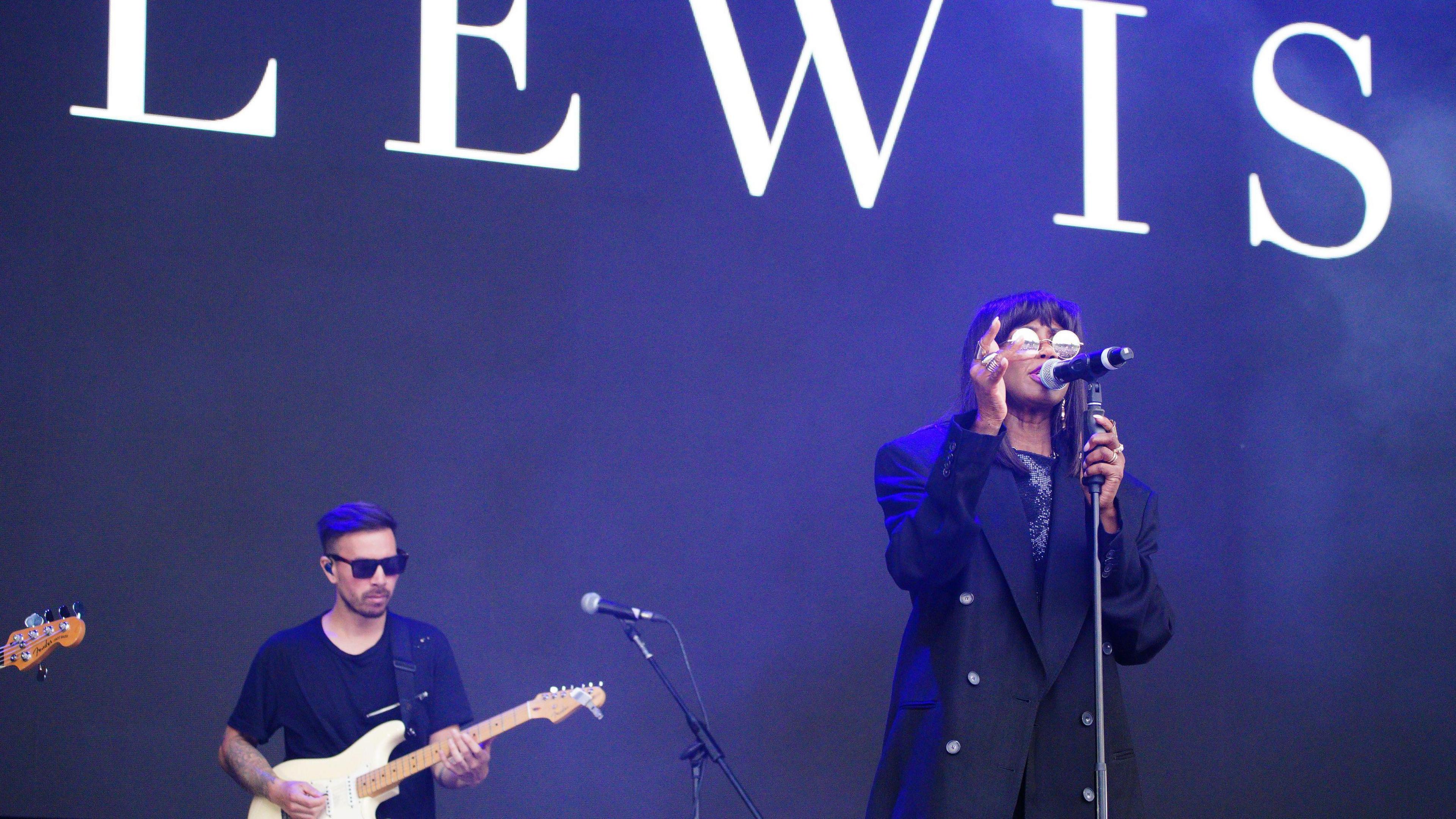 Shaznay Lewis singing on the left hand side of the screen at a mic stand - she is wearing distinctive round sun glasses. A guitarist is stood to the right of her. Her surname is up on the screen behind her.