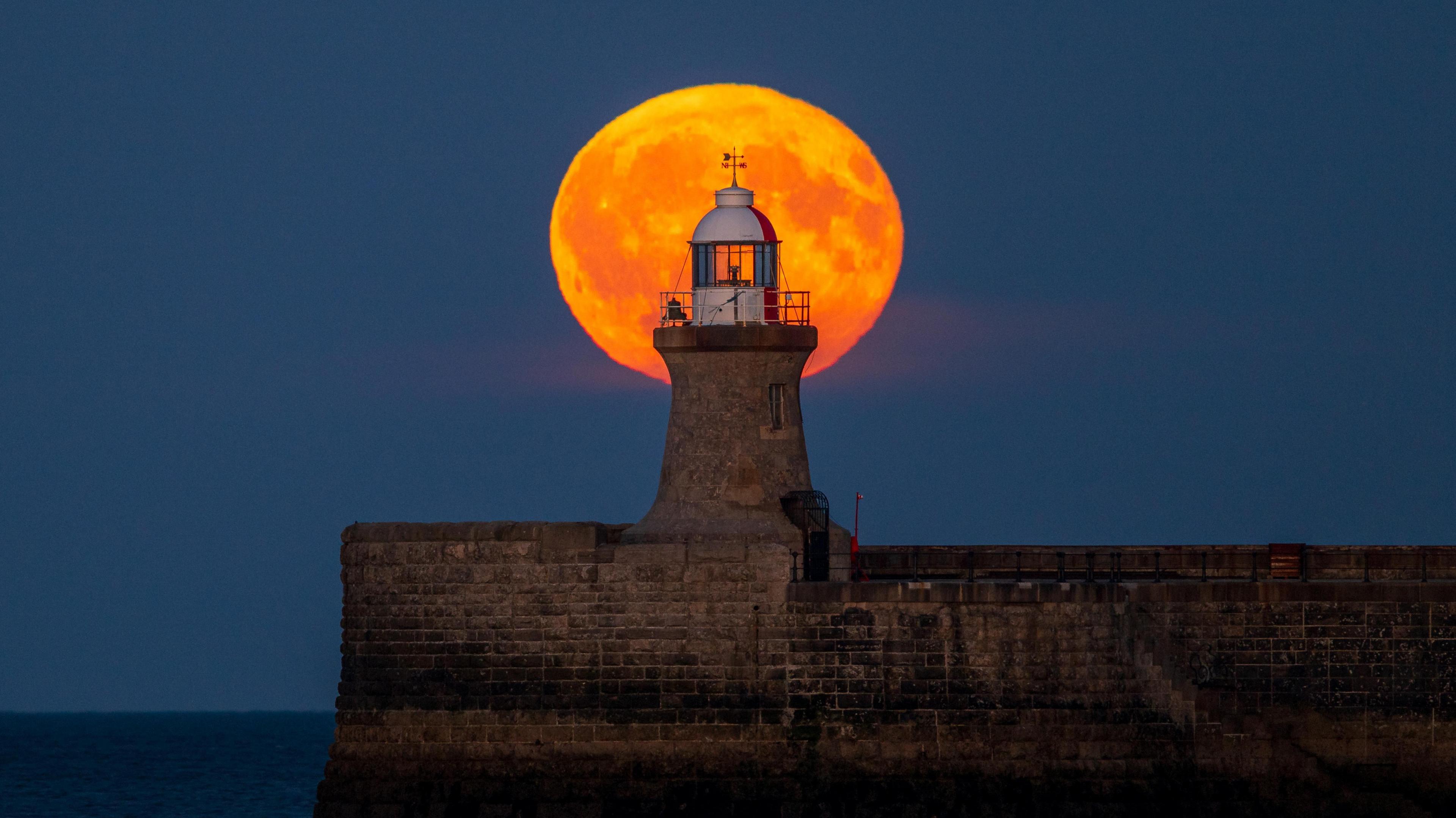 The moon is illuminated orange behind the red and white lighthouse at the end of the pier. 