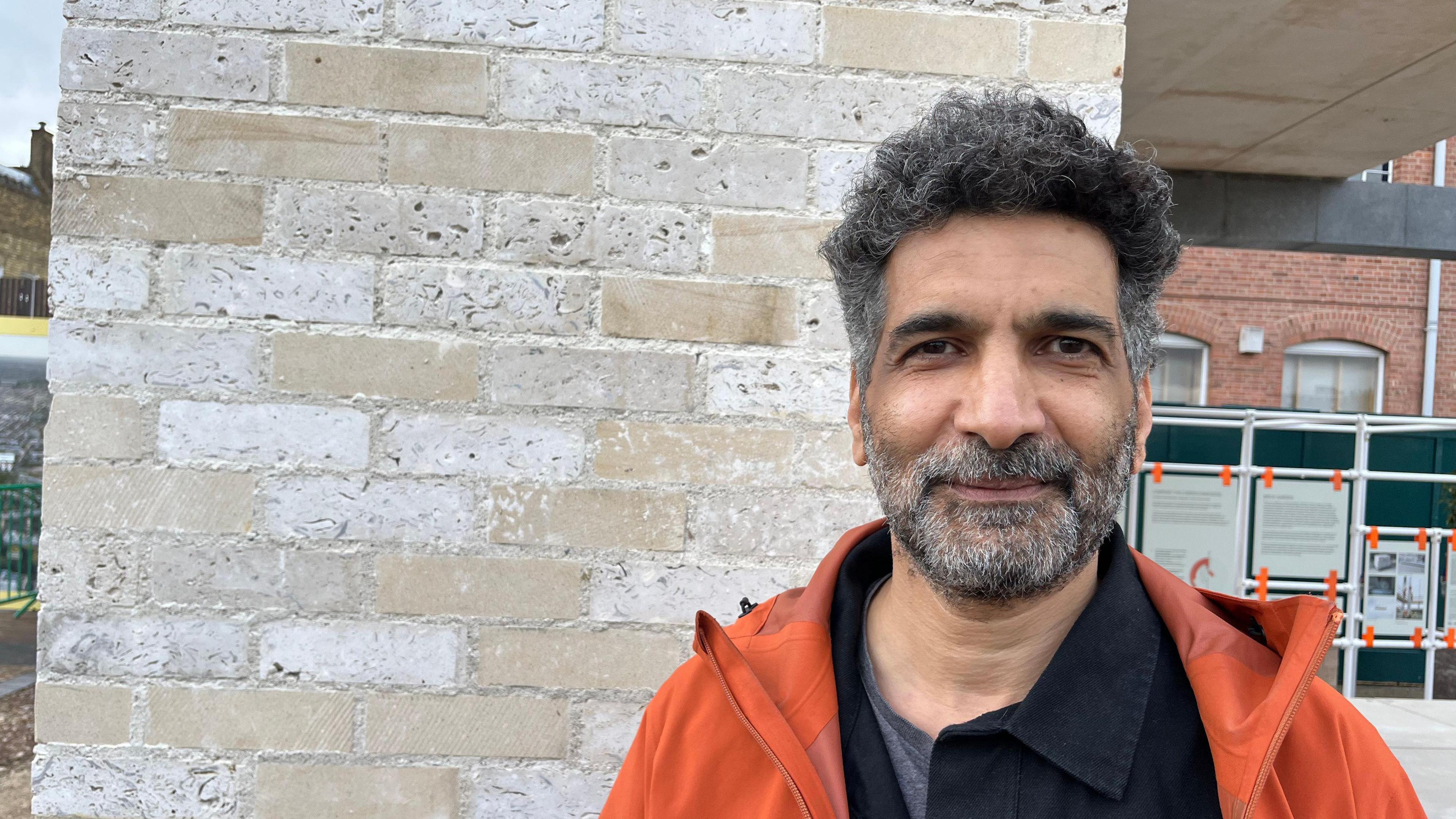 A man in an orange jacket looks at the camera. He is standing infront of a stone brick wall