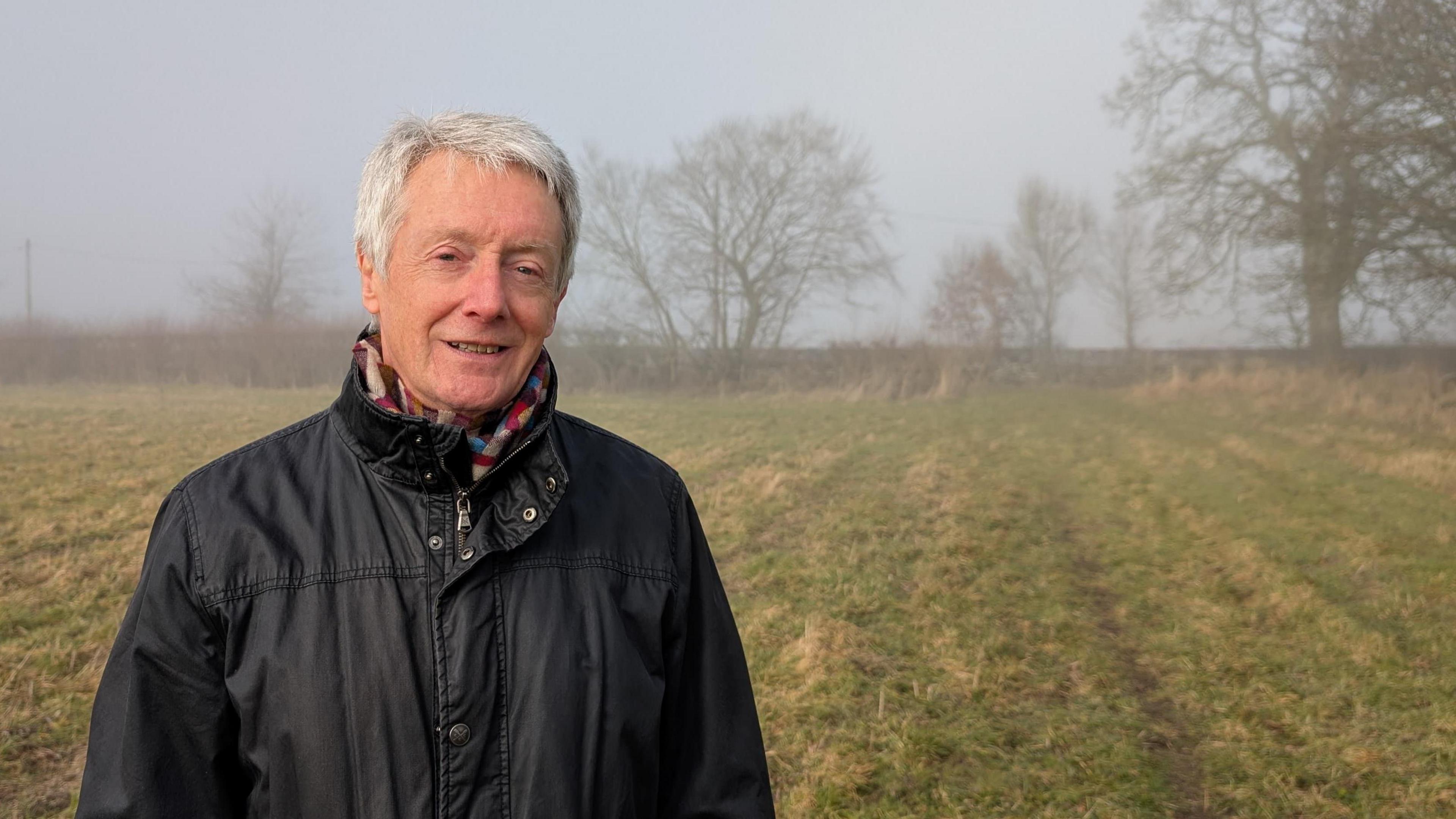 Nick Hayward smiles at the camera with the grassy field and some trees in the background. 