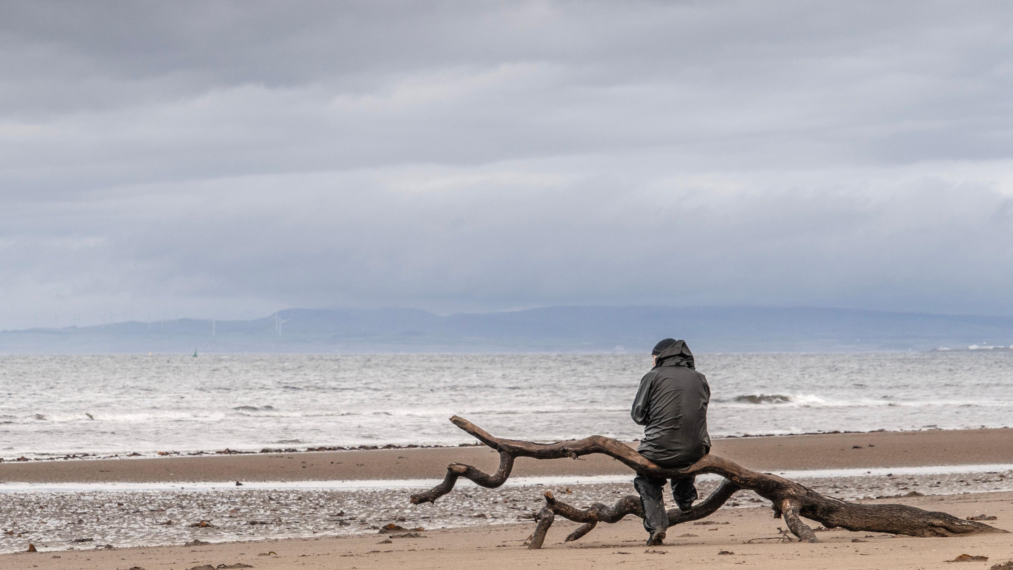 A man in a bunnet and wet weather gear sits on top of a large branch on a beach looking out to a grey and ominous sea and sky