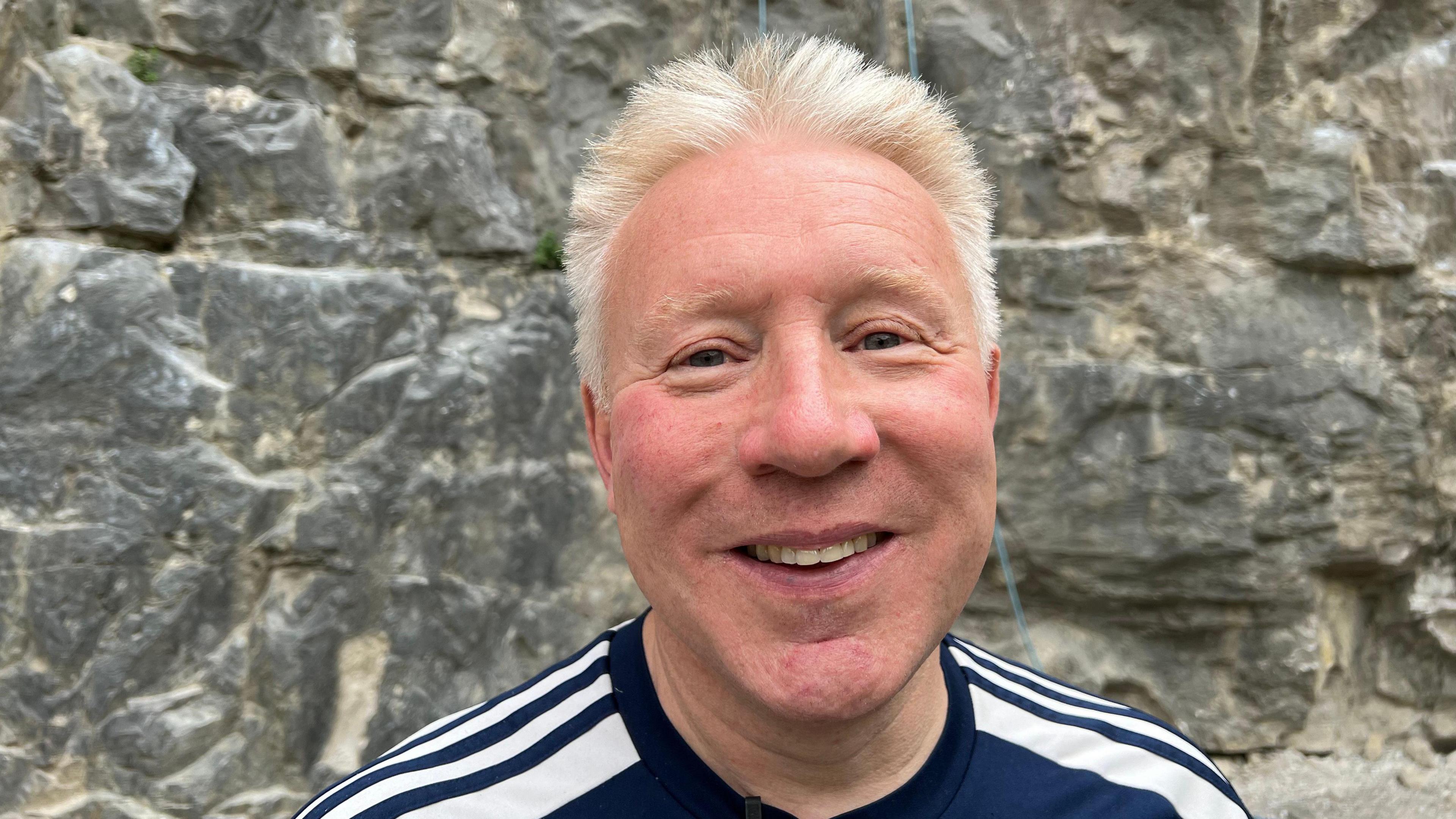 Older man with blond hair and a blue and white top in front of a rock face