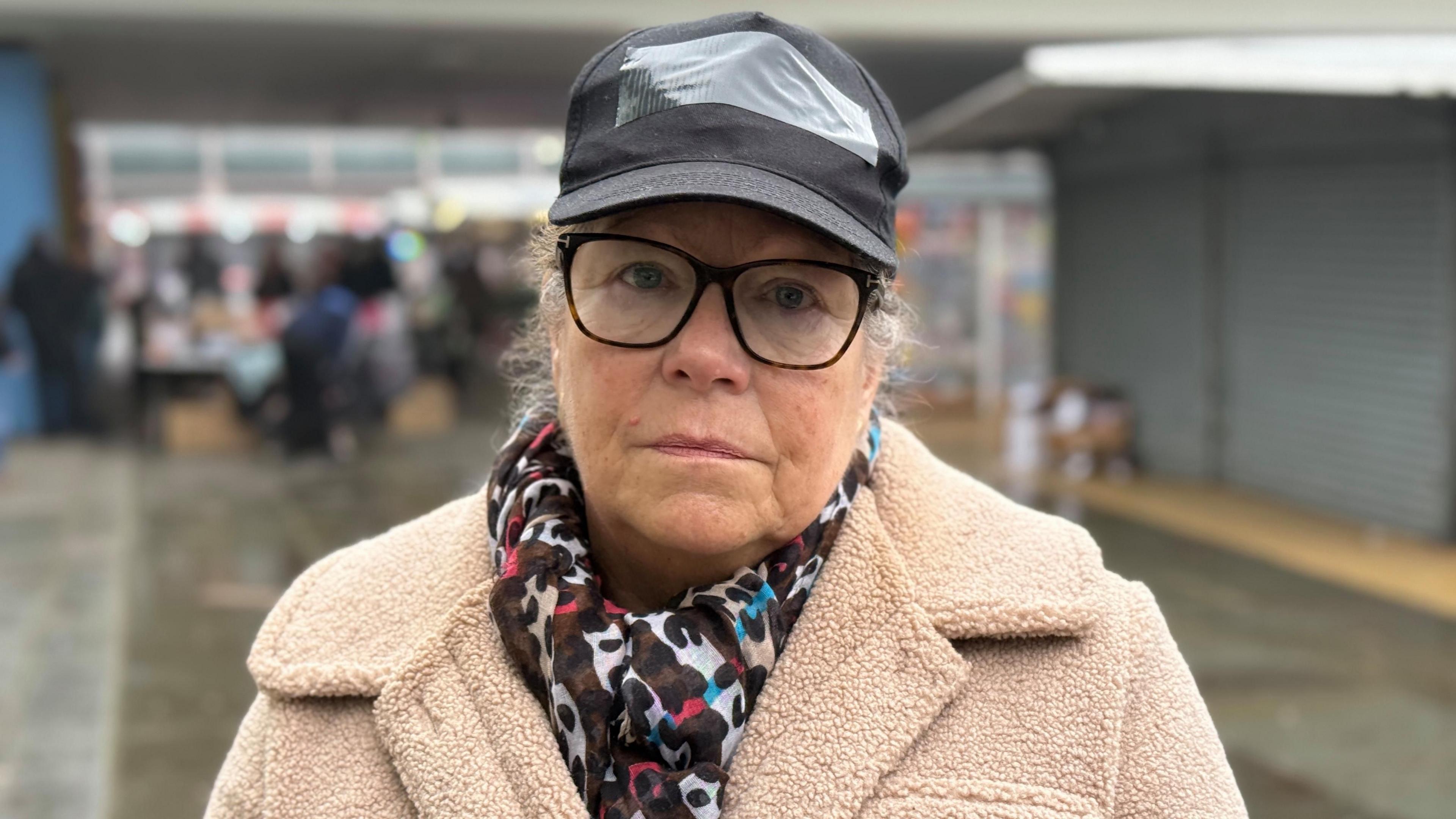 Maxine Herbert is wearing a cream coat, glasses and a black hat. She is standing in Milton Keynes Market.