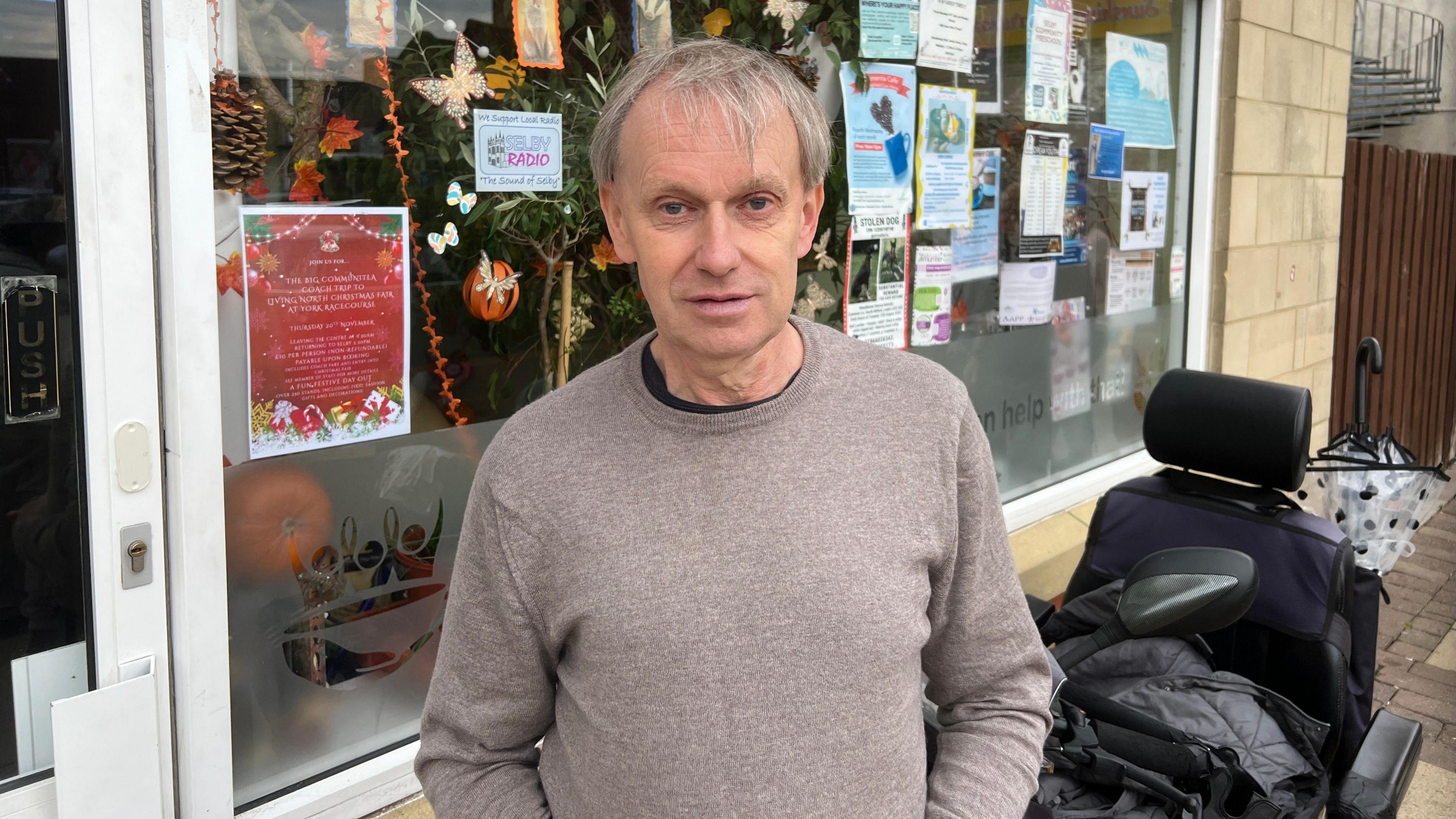 Neal Jackson is standing outside Selby mental health charity The Big Communi-Tea. He has grey hair and is wearing a light brown jumper. There are Halloween decorations in the window behind him.