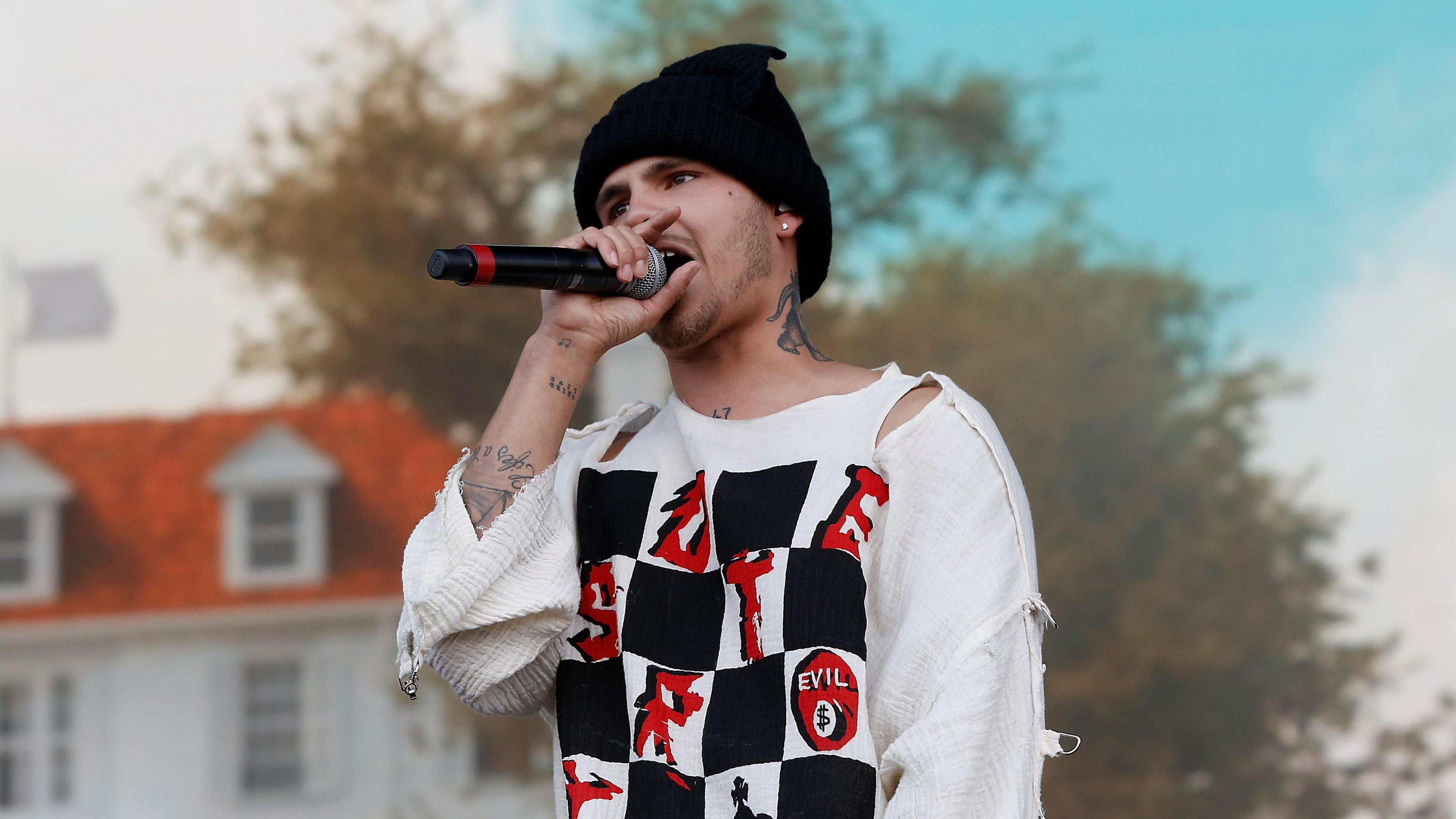 Slowthai performing with a microphone in his hand. He is wearing a black beanie hat and a ripped white shirt.