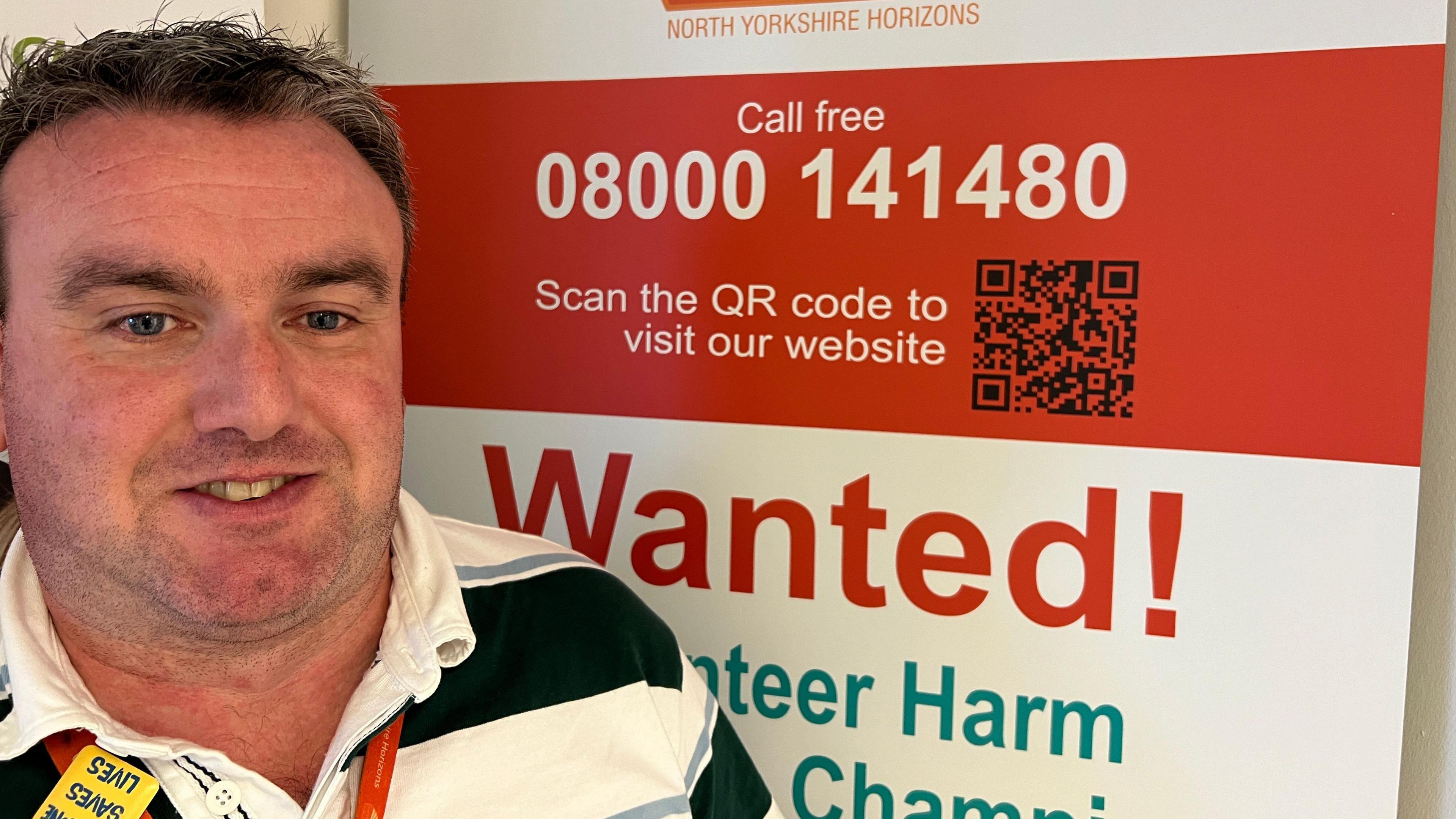 Kev Rafferty smiles towards the camera in front of a Horizons recruitment poster. He has dark hair and is wearing an open-neck black-and-white rugby shirt