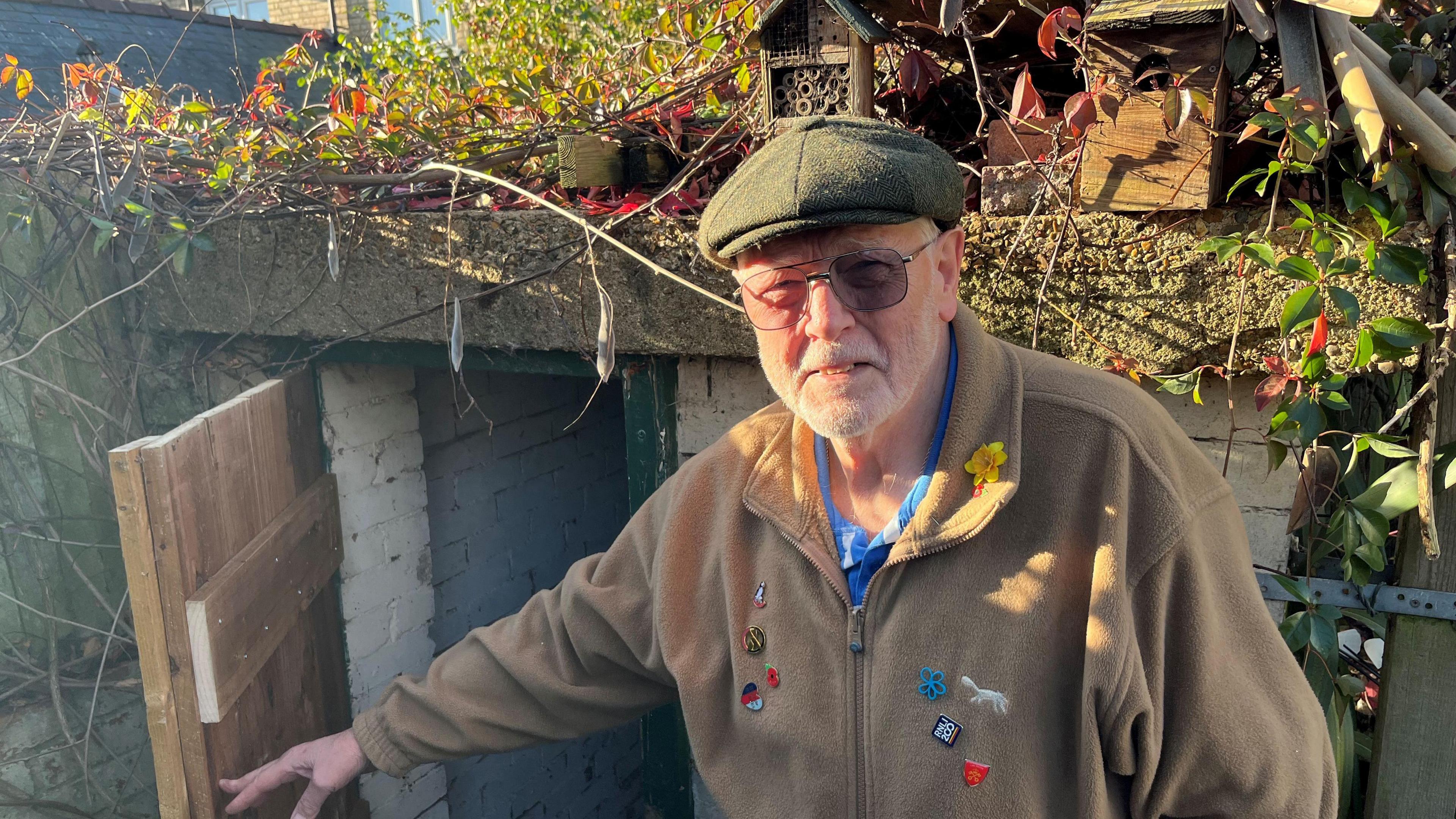 Colin Andrews has short grey hair and beard and has a green flat cap on his head. He has glasses with tinted lenses and is wearing a faun fleece over a blue, black and white shirt, he is holding the wooden door to the shelter open.