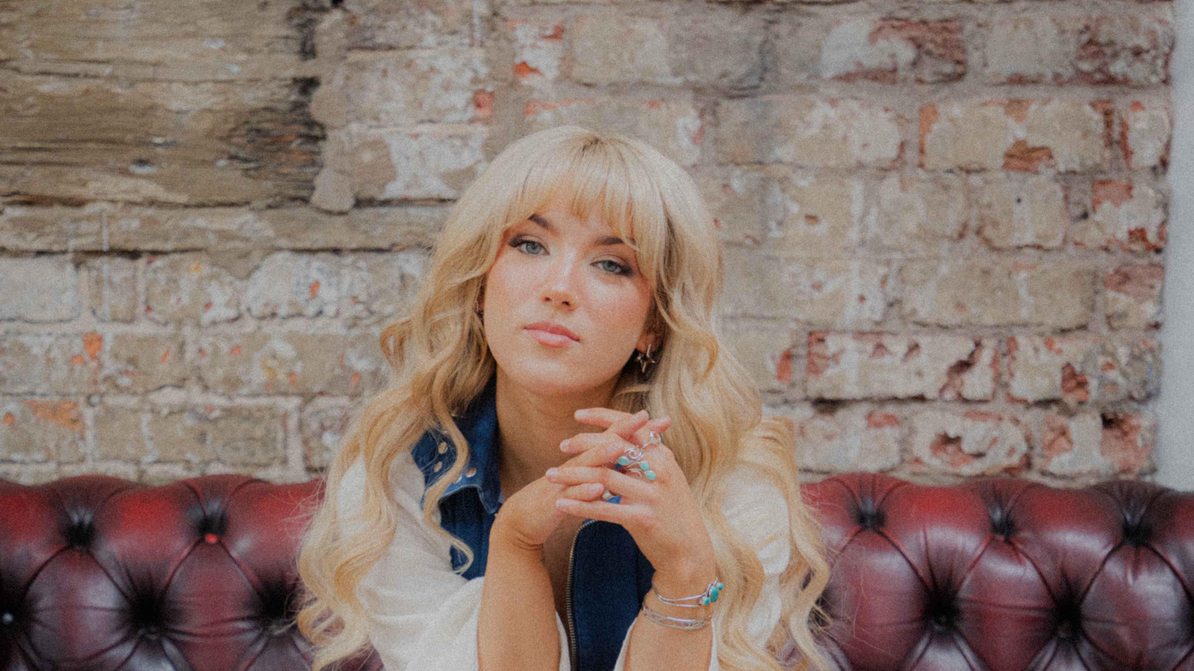 Rianne Downey looks into the camera while sitting on a red leather chair. She has a white and blue top on, and long blonde hair going past her shoulders.