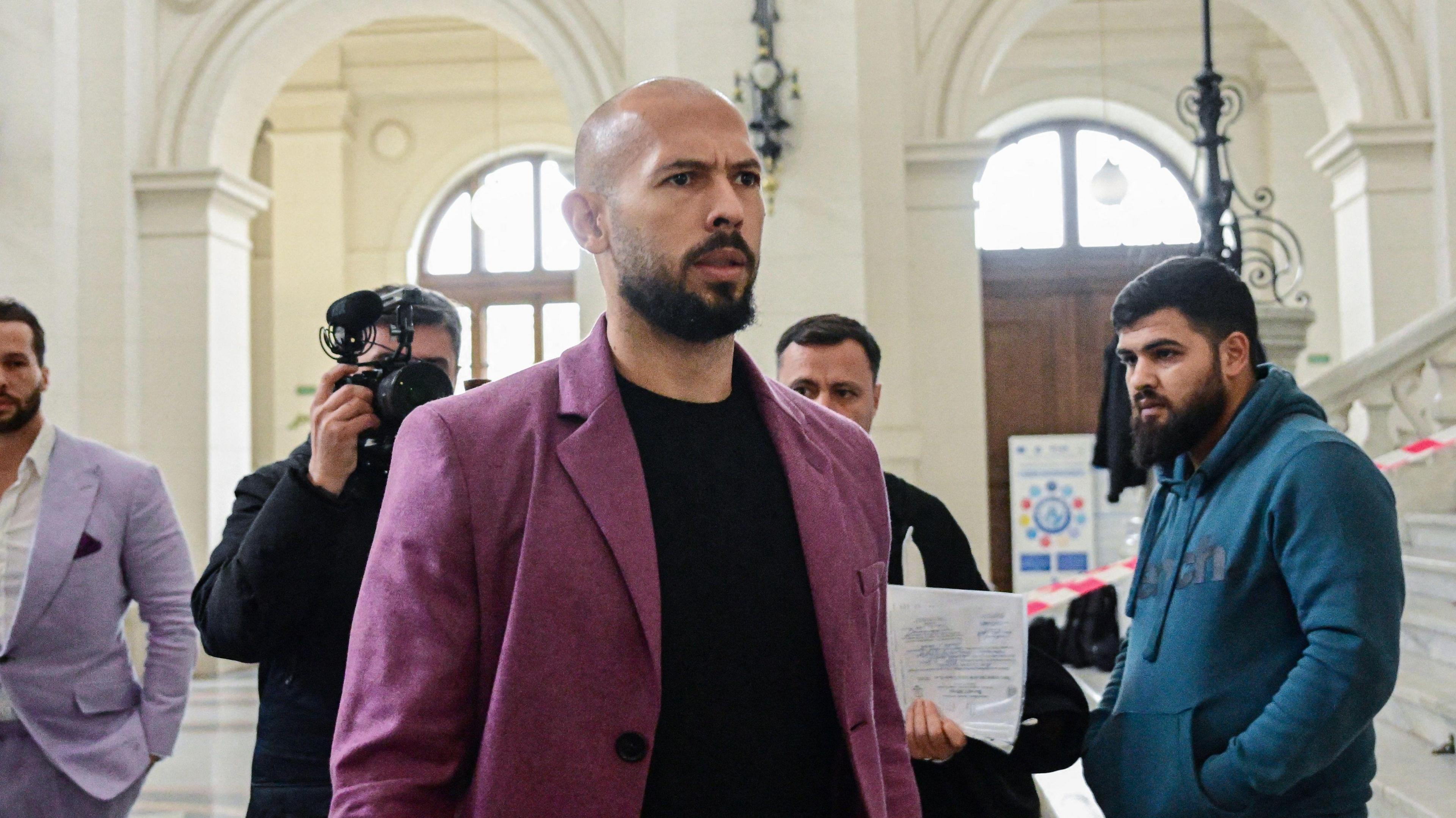 Andrew Tate wears a purple jacket as he walks inside a court in Bucharest, as a photographer behind him takes his picture 