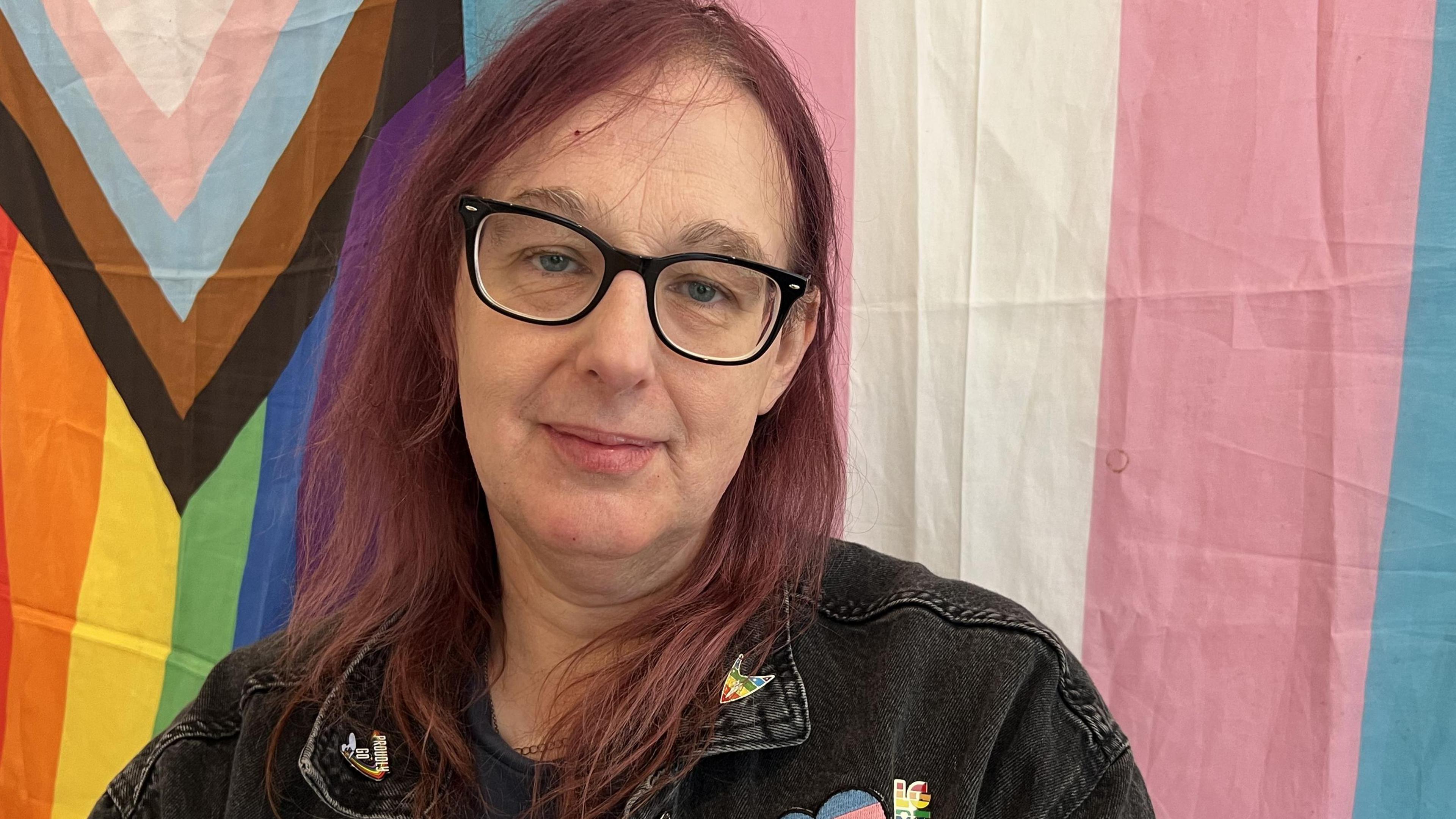 A woman with purple hair wearing glasses and a a black denim jacket with badges on. She is sat in front of a transgender and LGBTQI+ flag.