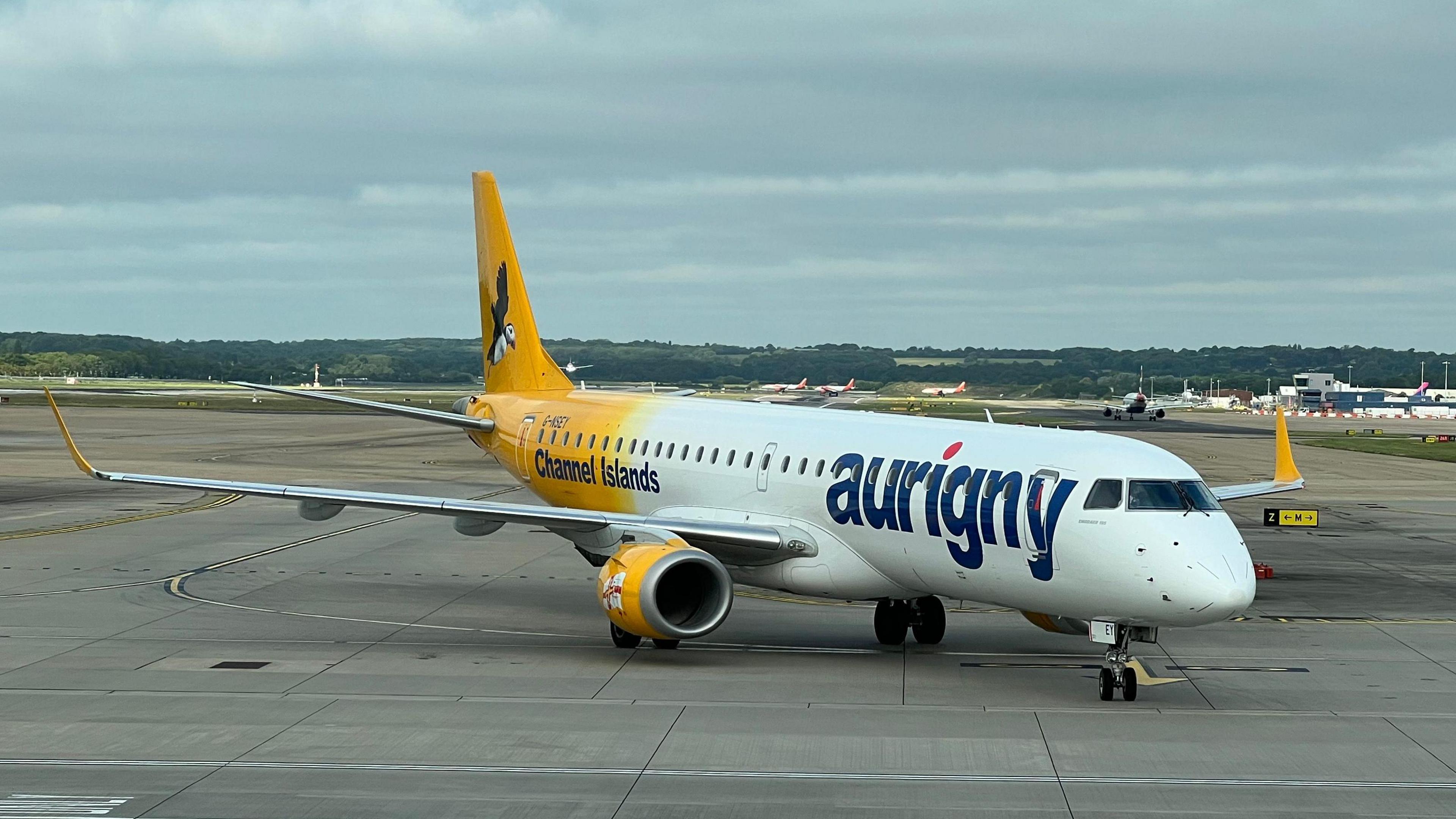 A shot of an Aurigny aeroplane at an airport. It is mainly white with the company branding on it with the back of it yellow. It has Aurigny and the Channel Islands labelled on it.