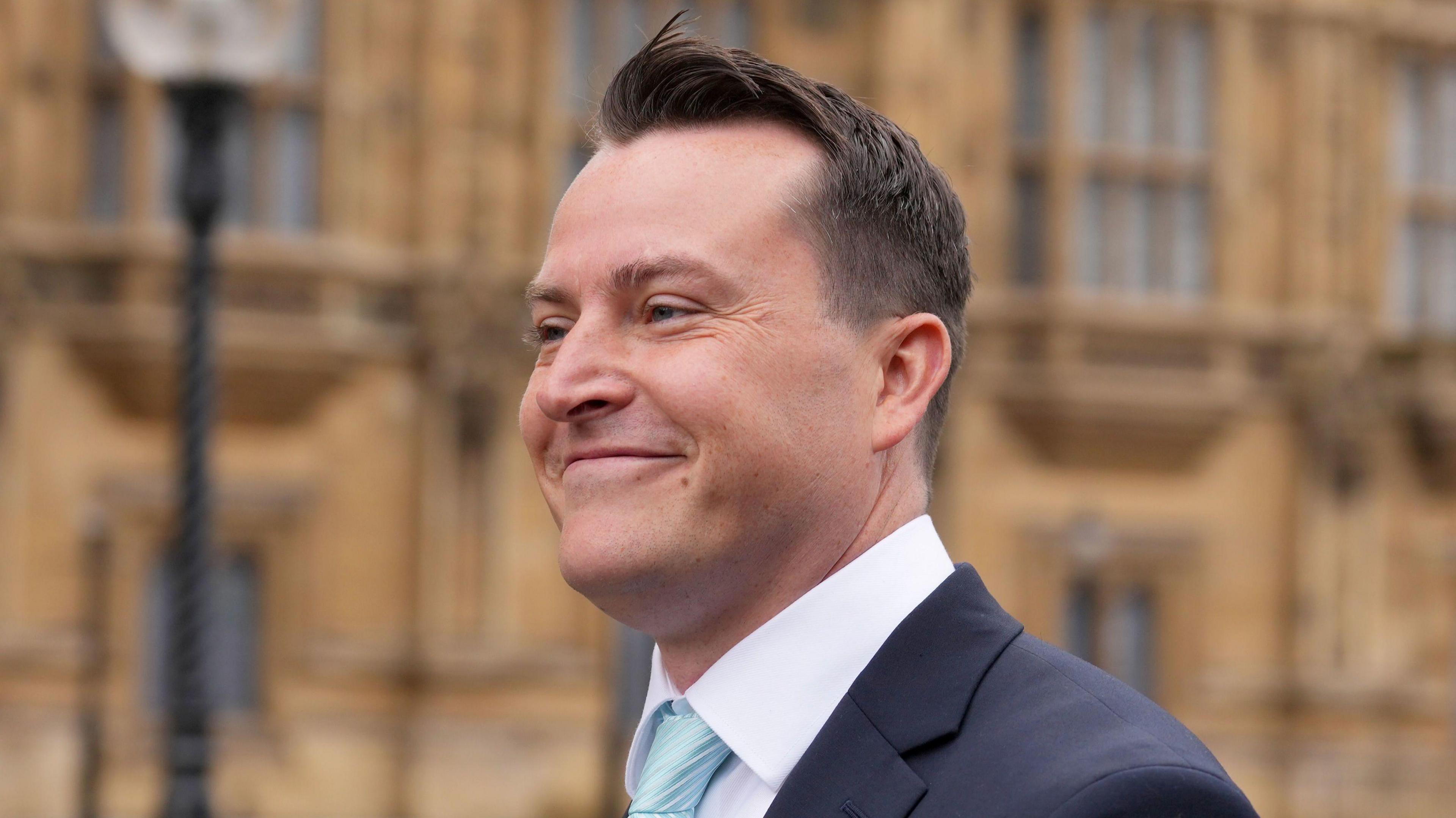 James McMurdock is wearing a blue tie, white shirt and navy blue suit jacket. He is side-on to the camera, smiling, and the Palace of Westminster appears to be out of focus in the background.