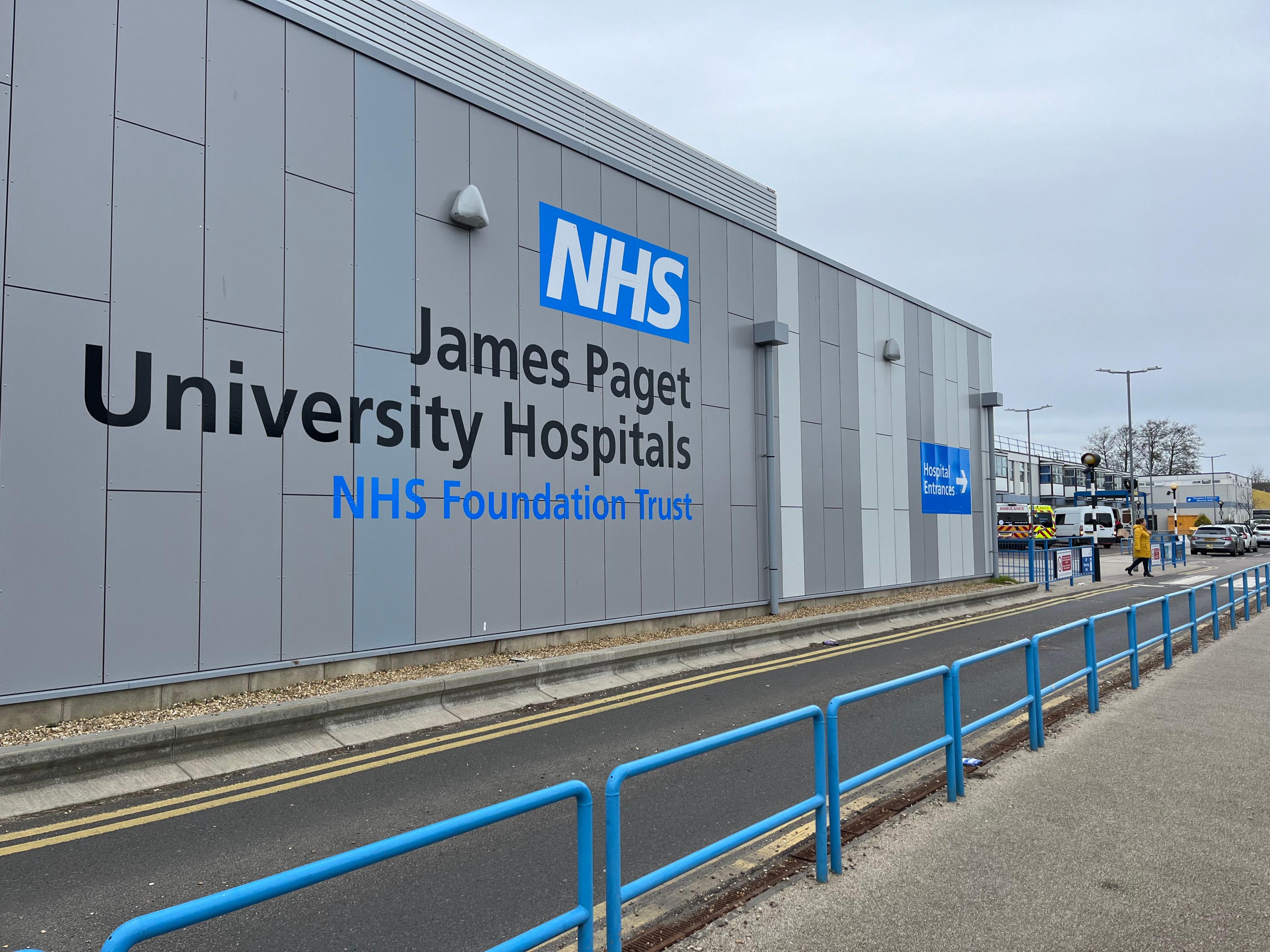 Exterior image of the James Paget Hospital in Gorleston. The large grey building has a blue guardrail around it and the name of the building is written on it in large letters